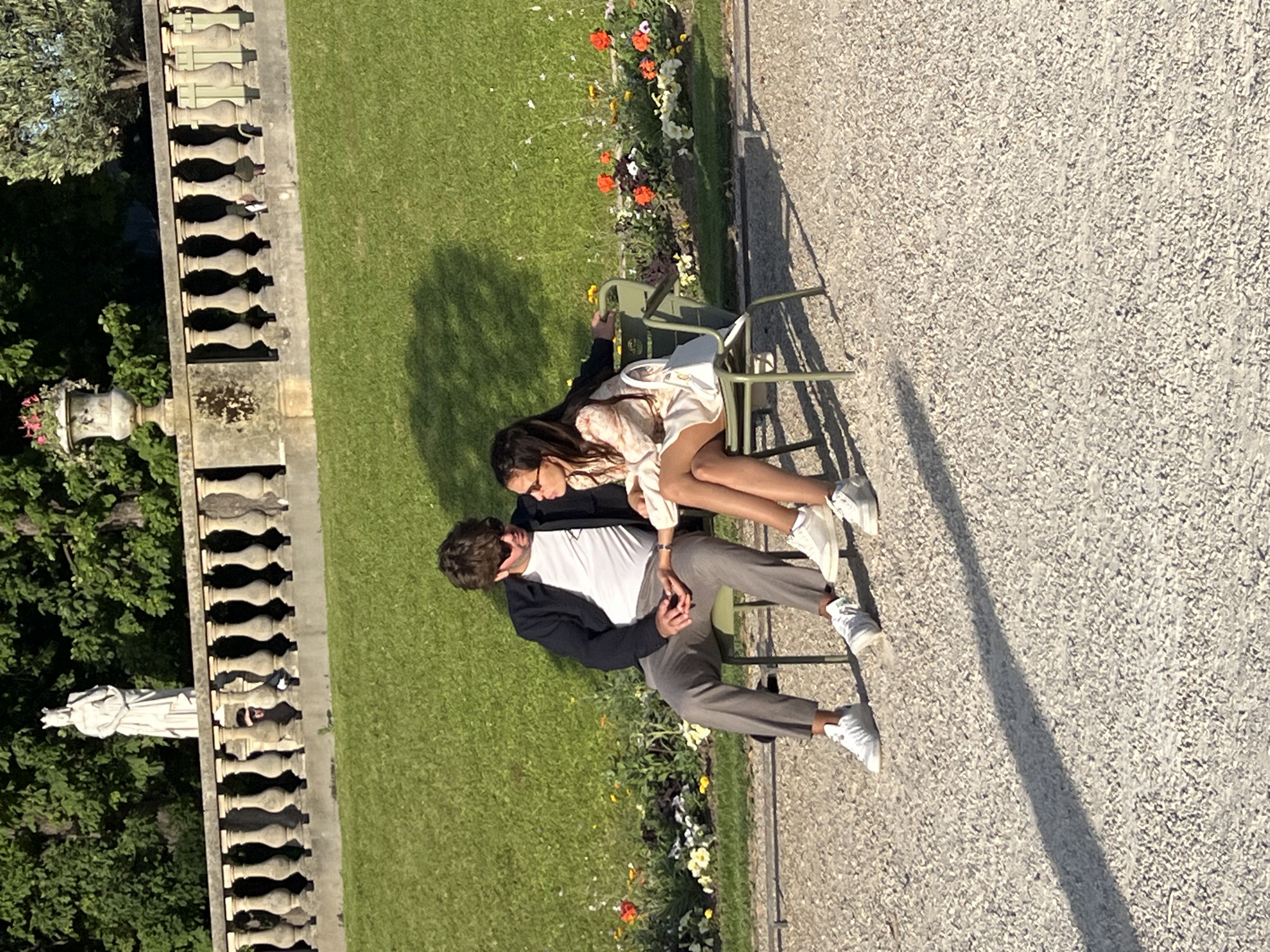 A couple sits on green chairs in a park, their legs touching, in Paris, France. The man, on the left, wears a dark jacket over a white t-shirt and gray pants. The woman, on the right, has on a patterned dress and glasses. A white purse rests on her lap. They appear to be looking at each other. In the background, a stone balustrade lines a grassy slope dotted with flowers. A white statue stands atop the slope. The ground in the foreground is covered in gravel.