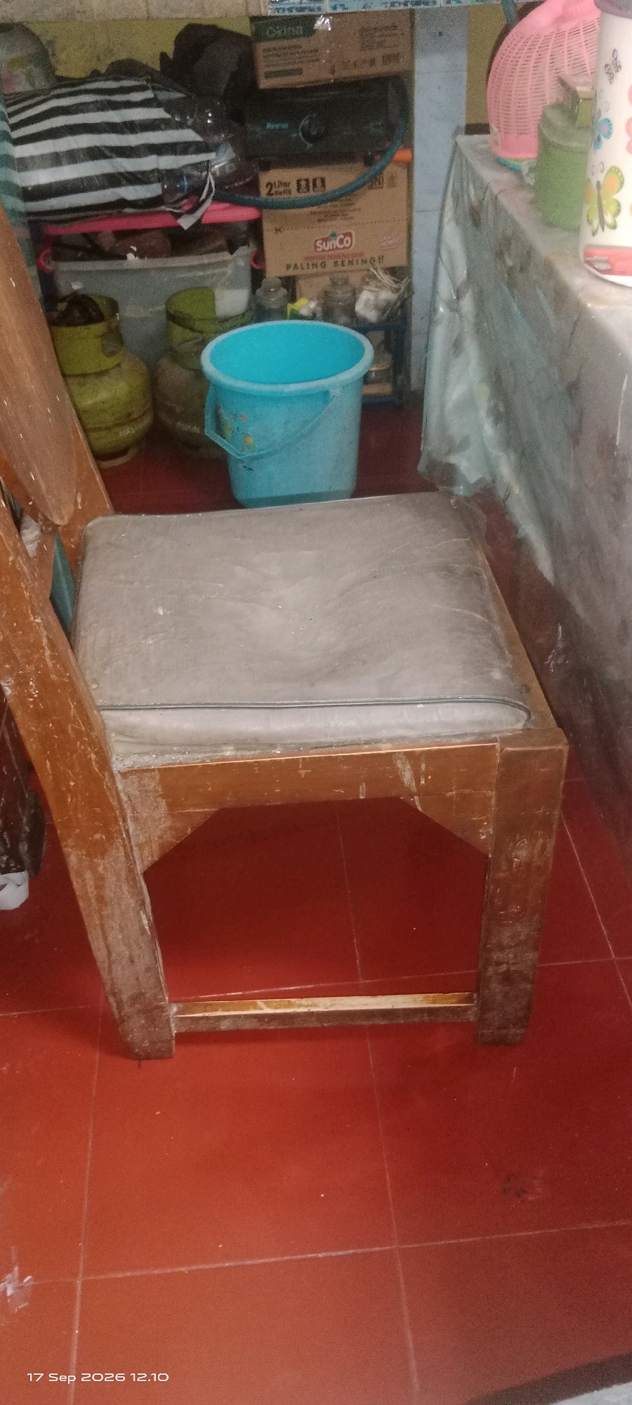 The image shows a cluttered corner of what appears to be a storage area or informal living space, likely in Klaten, Indonesia. In the foreground, a wooden chair with a worn, grey upholstered seat is positioned on a red tiled floor. The chair is made of dark wood and shows signs of age and use, with scuff marks and faded areas.
Behind the chair, to the left, are several items stacked against a wall. These include what appear to be two large green propane tanks, a large black and white striped bag, and a blue plastic bucket. Further back and to the right, there are boxes with various labels, including one with "SunCo" prominently displayed. There are also some glass jars and other miscellaneous items.
To the far right, a table is partially visible, covered with a light-colored plastic sheet. On top of this table are household items, including a pink plastic basket and what looks like a pink and white trash bin with a butterfly design. The overall impression is one of a functional, lived-in space that is not meticulously organized. The presence of propane tanks and the general clutter suggest a residential or small business environment. The timestamp "17 Sep 2026 12.10" is visible in the bottom left corner of the image.