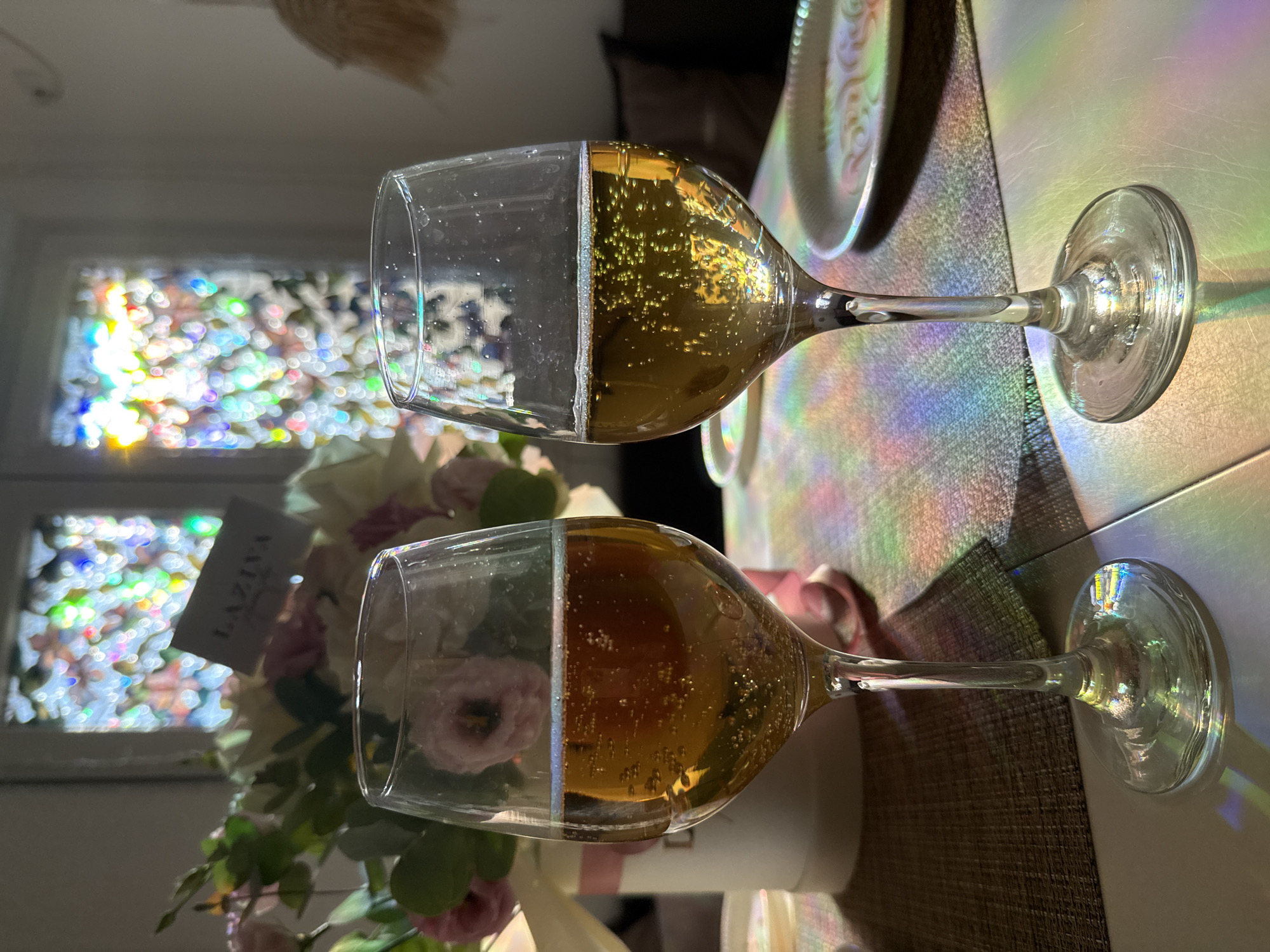 Two wine glasses filled with a golden-brown beverage, possibly champagne or cider, are placed on a table. The glasses are clear and have a slight shimmer, with small bubbles visible in the liquid. In the background, a bouquet of pink and white flowers sits in a white vase, with a small card bearing the word "LAZIVA" partially obscured by the flowers. Behind the flowers, a window or screen displays a colorful, bokeh effect of heart shapes. The table is covered with a textured placemat, and there are plates visible on either side of the glasses. Rainbow-like light refractions are cast across the table and on the glasses, indicating a light source creating these effects. The scene appears to be set indoors, possibly for a celebration or a special occasion. The location is Unknown, Unknown.