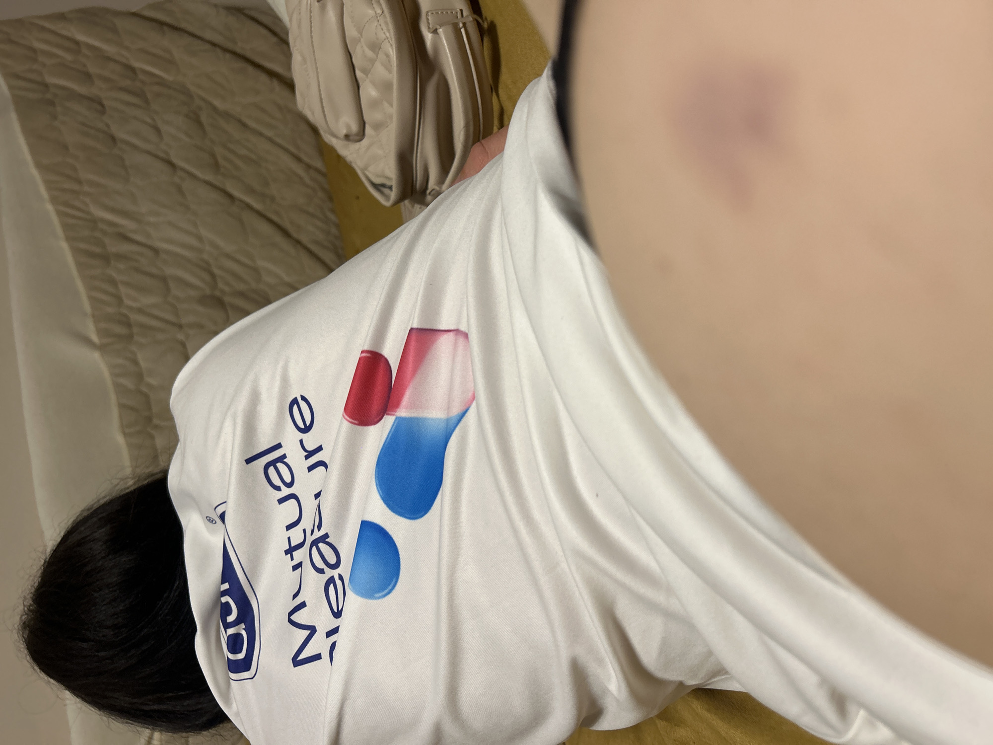 A close-up indoor shot shows a person with dark hair wearing a white t-shirt with a "Mutual Measure" logo. The person is lying on a quilted, light brown surface that appears to be bedding. A beige handbag is partially visible near the person's shoulder. A purplish bruise is visible on bare skin near the edge of the frame. No context indicates the location is Burgas, Bulgaria.