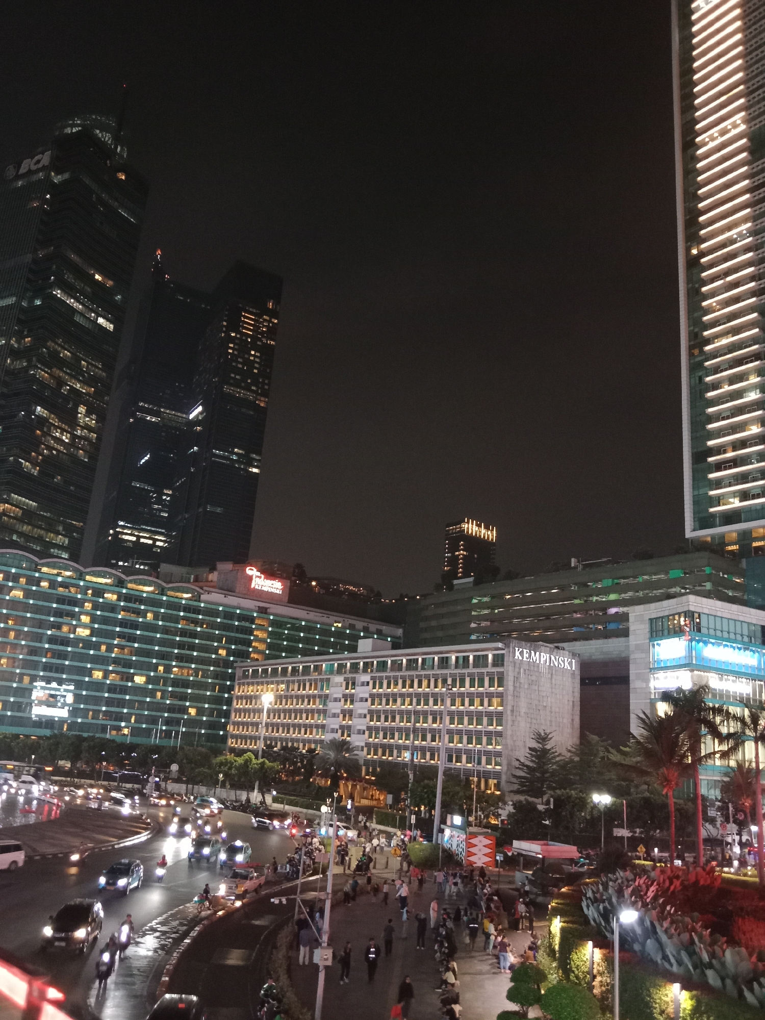 The image depicts a nighttime street scene in Central Jakarta, Indonesia, likely in the vicinity of the Bundaran HI intersection. The foreground shows a busy road with multiple lanes of traffic, illuminated by streetlights and the headlights and taillights of vehicles including cars, motorcycles, and a few pedestrians are visible on the sidewalks and crossing the street. The road surface appears wet, suggesting recent rain.
Dominating the background are several tall, modern skyscrapers, brightly lit from within and along their facades. One prominent building features the "BCA" logo. Another tall, slender building on the right is characterized by its vertical lines of illumination. In the mid-ground, a lower, wider building with a grid-like facade is visible, bearing the prominent signage "KEMPINSKI." This building also features many illuminated windows and some landscaping with palm trees. The overall impression is of a vibrant, bustling urban environment at night.