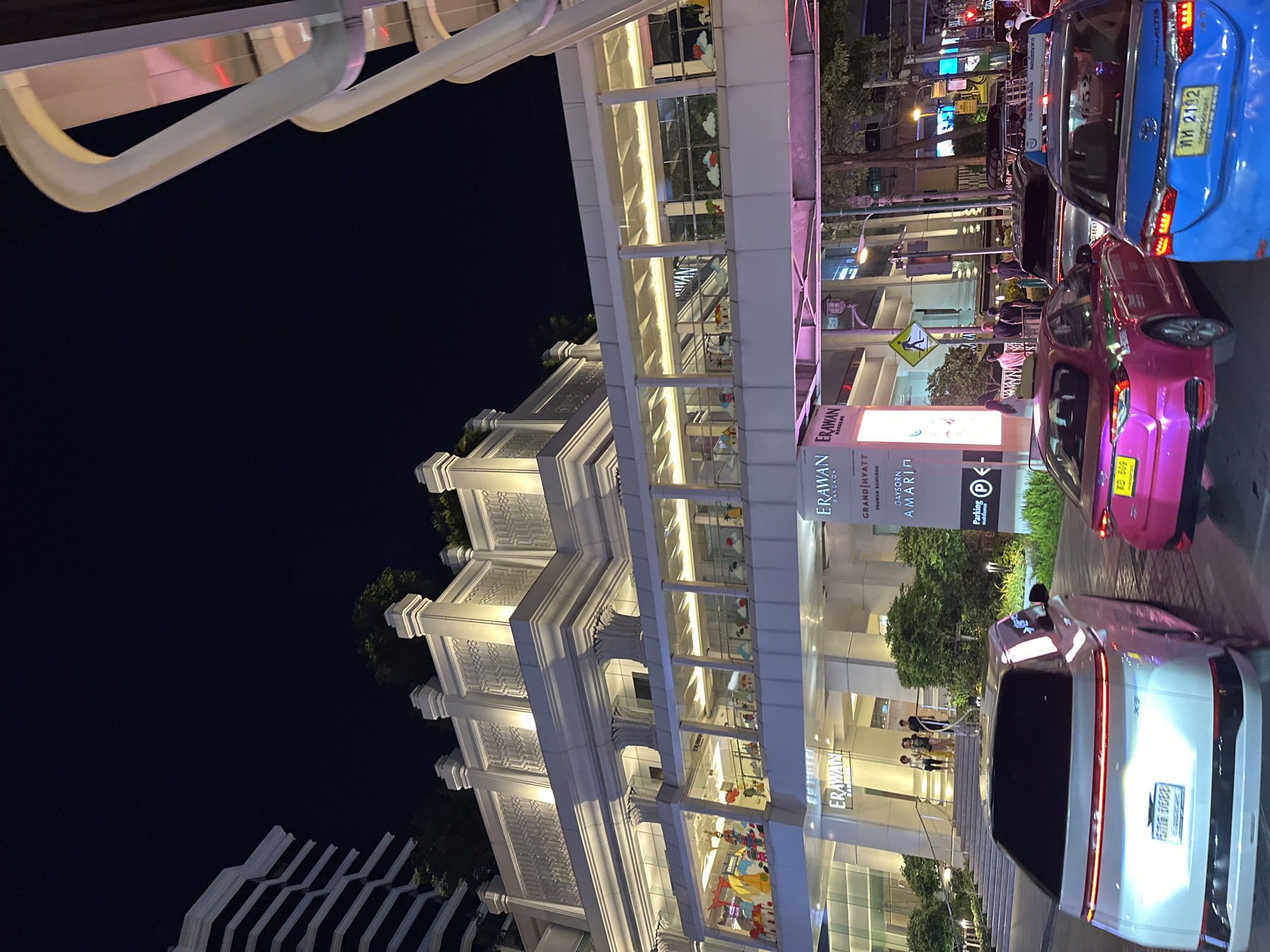 This is a nighttime, street-level view of the Erawan Bangkok shopping center in Bangkok, Thailand. The architectural style is ornate with columns and lighted windows. Attached to the building is a pedestrian overpass. Street traffic, including a blue Toyota, pink car, and a white vehicle with a visible license plate, is present. A sign displays the names of businesses, including Erawan Bangkok, Grand Hyatt Erawan Bangkok, Gaysorn Amarin, and a parking symbol.
