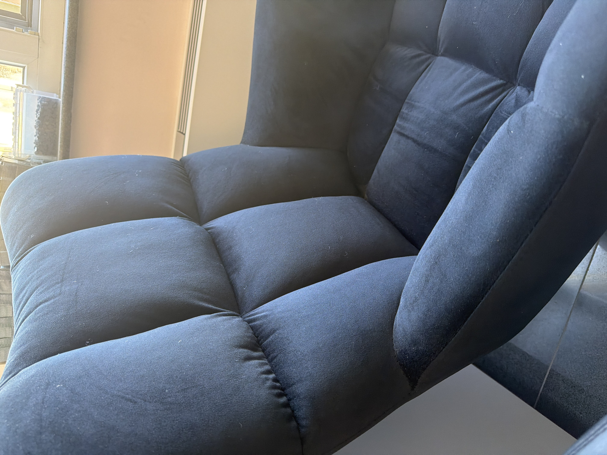 The image shows a close-up of a dark blue upholstered armchair. The chair has a tufted back with visible stitching and padded sections. The armrest is also upholstered and appears to be soft. The chair is positioned indoors, with a hint of a window and a radiator visible in the background. The lighting suggests it is daytime, with sunlight casting shadows on the chair. There is no indication of the city of Dolno Ablanovo, Bulgaria in the image. The scene is static and focuses solely on the armchair.