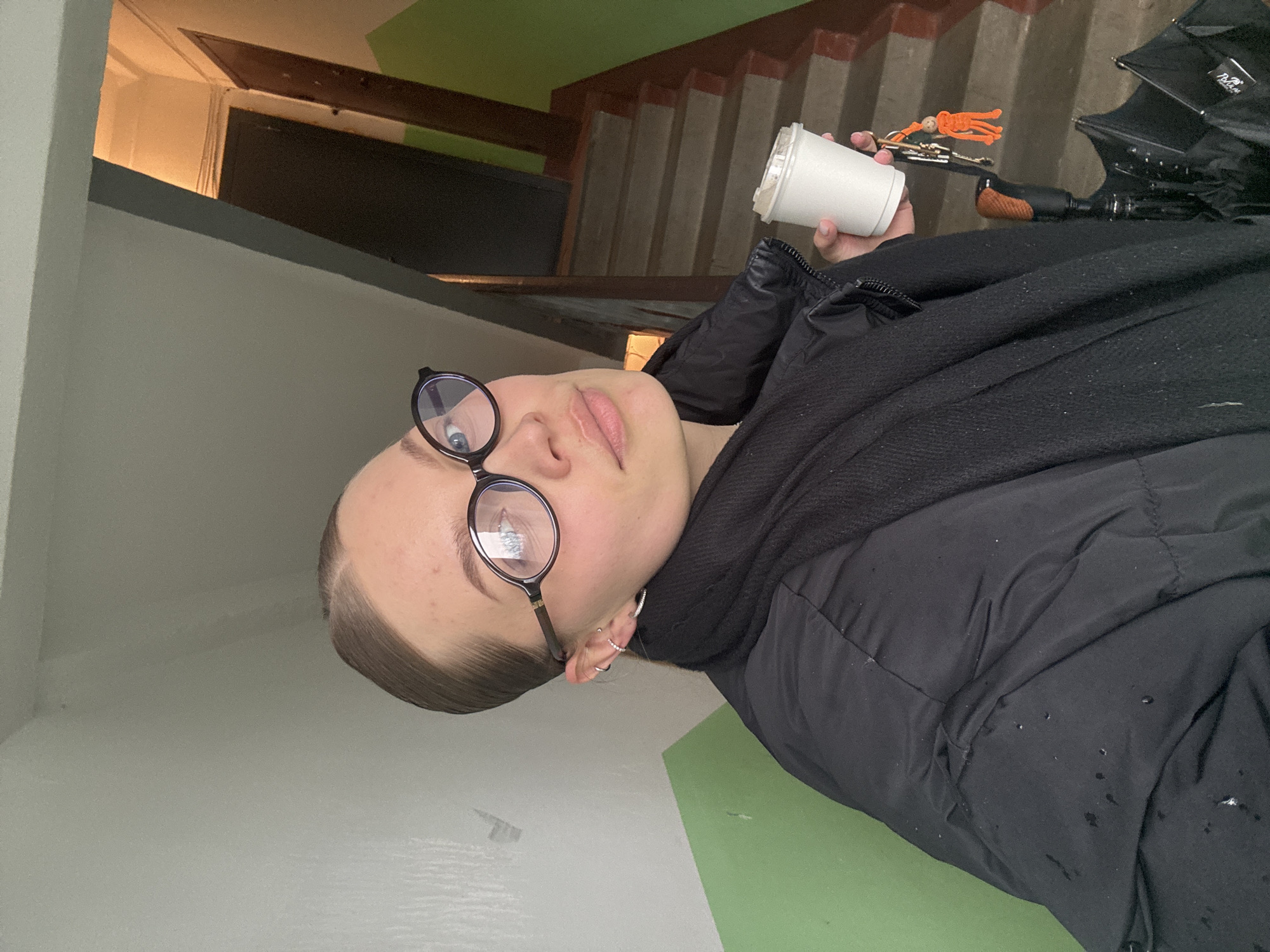 A person is holding a coffee cup and keys, standing on a landing of a staircase with an umbrella nearby. The person is wearing glasses, a black puffer jacket, and a scarf. The staircase has concrete steps with red and white risers. The landing is painted white and green. The location is implied to be in the city of Chaikovsky, Russia.