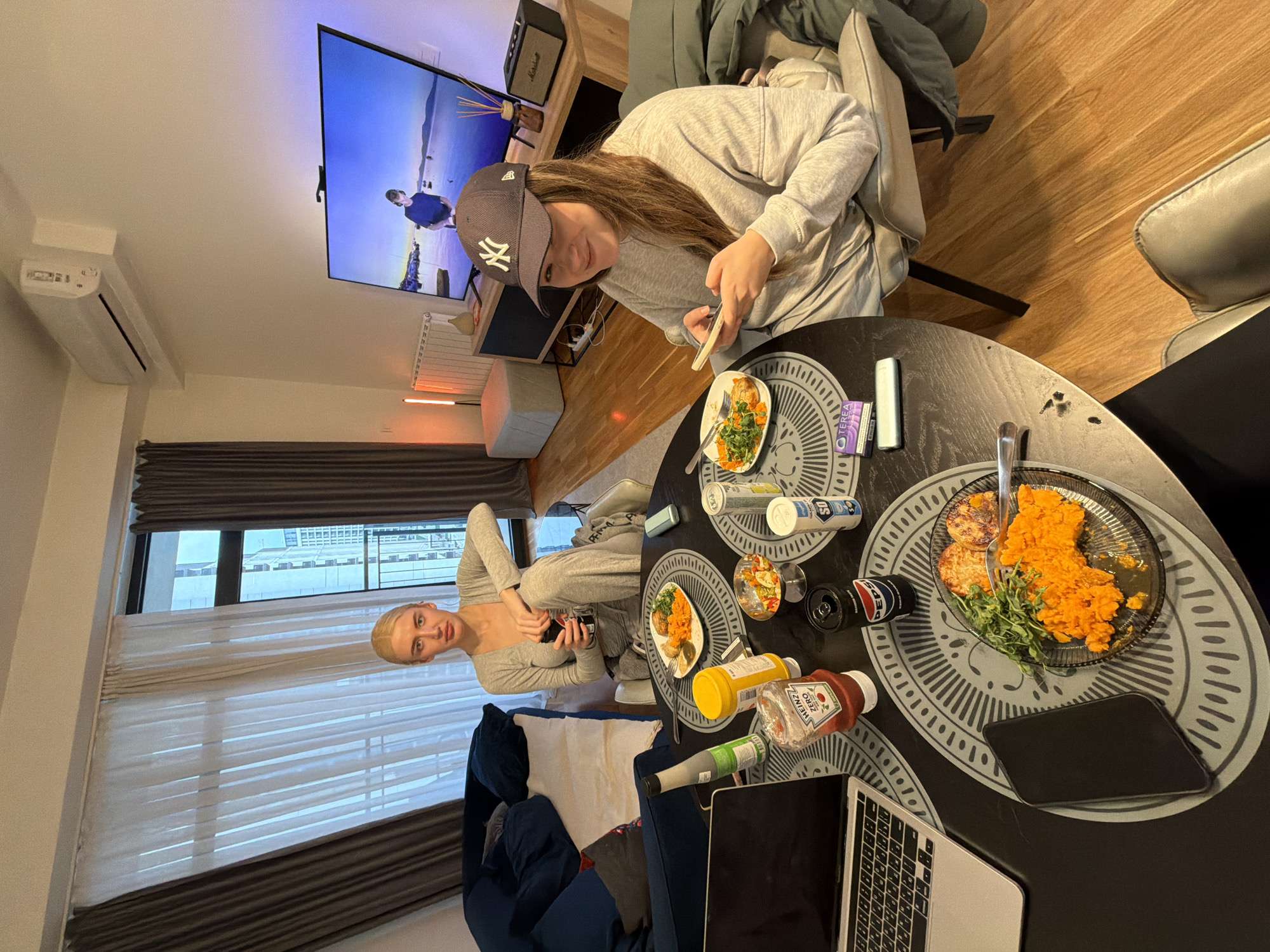 Two young women are in an apartment. One woman is seated at a round, black table, wearing a grey sweatsuit and a New York Yankees baseball cap. She is looking at a mobile phone and holding chopsticks. The other woman is seated on a blue sofa by a window, also wearing a grey sweatsuit and holding a mobile phone. The table has plates of food, condiments, beverages, and a laptop. A large TV screen is visible on the wall behind the woman at the table. The location is Belgrade, Serbia, as indicated by the window view and the "TEREA" cigarettes visible on the table.