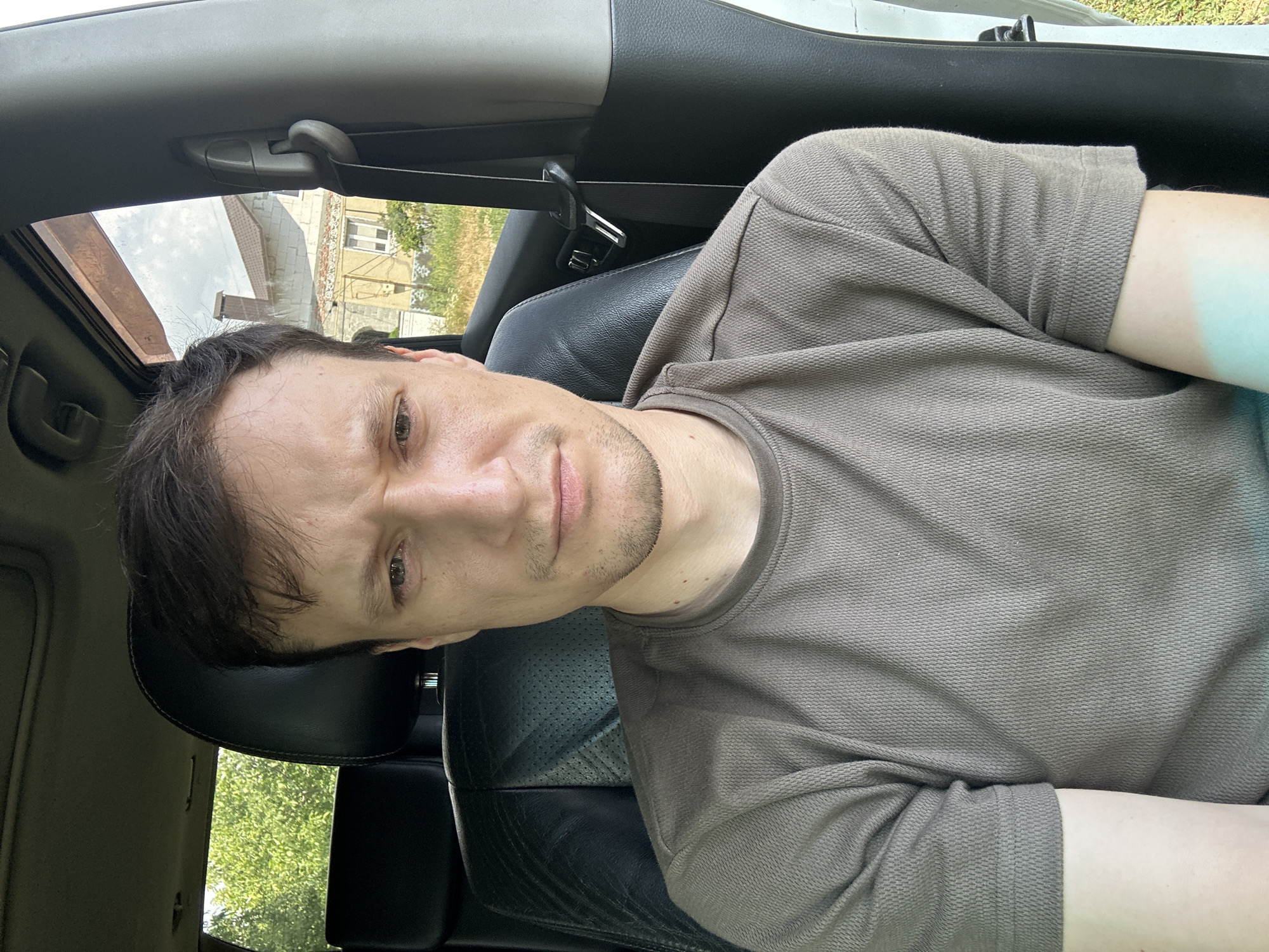 A man with brown hair and a faint stubble is seated in the driver's seat of a car. He is wearing a brown, textured t-shirt. His facial expression is a grimace, with furrowed brows and a downturned mouth. The car interior features black leather seats and a seatbelt. Outside the car, through the windows, a residential building with a light-colored facade and a dark-tiled roof is visible, along with green trees and dry grass. The scene appears to be outdoors, possibly in a rural or suburban setting. The location is identified as Makiv, Ukraine.