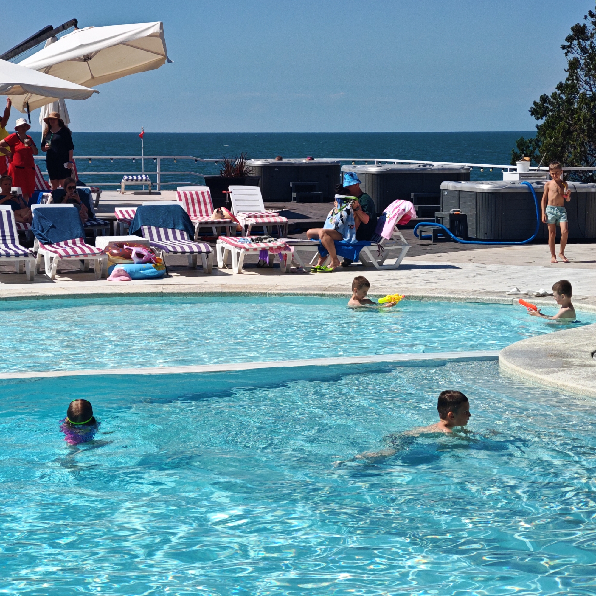 This is an outdoor medium shot of a group of people at a pool on a sunny day. In the foreground, a young boy in the pool is holding a red water gun. To his left, another young boy in the pool holds a yellow water gun. Further back in the pool, a young girl with green goggles and a purple swimsuit is swimming.
In the middle ground, there are several lounge chairs with striped cushions, some with towels draped over them. A man wearing a blue hat is sitting on a lounge chair, reading a book. Beside him, a young boy is standing, holding a red drink. In the background, there are two large hot tubs and a railing overlooking the ocean.
Further back, several people are standing on a deck. One woman in a red dress and white hat is raising her hand, seemingly waving. Another woman in a black dress and a straw hat is holding a drink. The scene appears to be at a resort or hotel, with the ocean visible in the distance. The weather is clear and sunny, suggesting it is daytime. There is no visible text in the image.