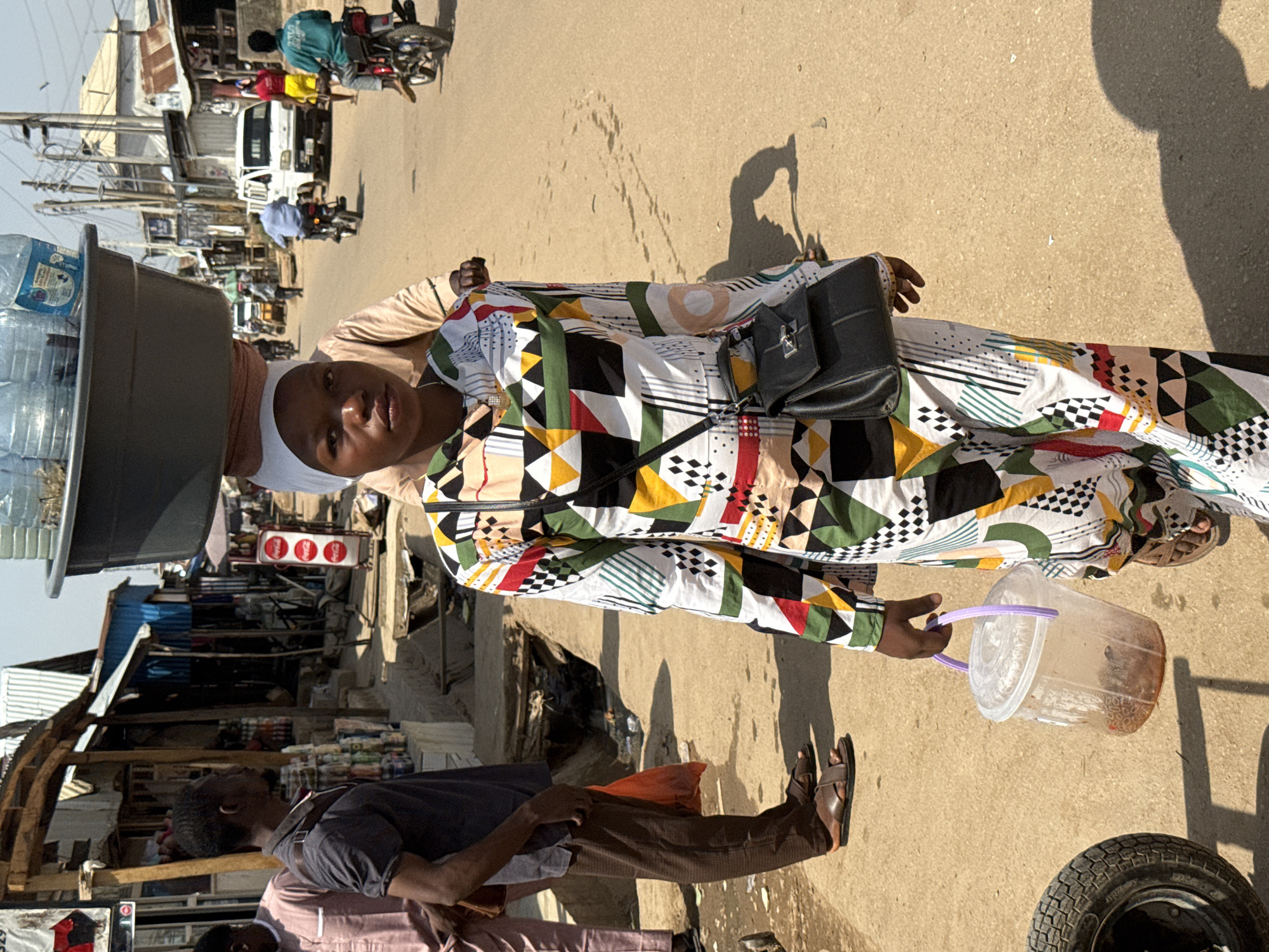 The image captures a vibrant street scene in what appears to be a market or commercial area in Abuja, Nigeria, during a bright, sunny day, indicated by the strong shadows cast on the ground.
The central subject is a young woman, likely a street vendor, facing the camera with a calm, neutral expression. She carries a large, dark basin filled with plastic bottles of water on her head. She is dressed in a striking long-sleeved dress featuring a multi-colored geometric pattern in white, black, green, yellow, red, and brown. A white head covering completes her attire, and she wears a black shoulder bag. In her left hand, she holds a transparent plastic bucket with a purple handle, containing a reddish-brown substance, possibly another item for sale.
In the mid-ground to her left, a man in a dark blue shirt, brown trousers, and brown sandals stands, looking towards the right. Further into the background, other individuals are visible, some on motorbikes ("okadas"), and others walking, indicating a busy thoroughfare.
The setting consists of unpaved ground, and the background reveals various makeshift market stalls or shops with corrugated metal roofs and basic structures. Electrical poles and wires crisscross the sky. Visible text includes "Coca-Cola" logos on three red circular signs in the background and faint branding ("BIG WATER") on some of the water bottles on the woman's head. A single tire lies on the ground in the bottom right corner. The overall scene depicts typical daily commerce and life in a bustling Nigerian street market.