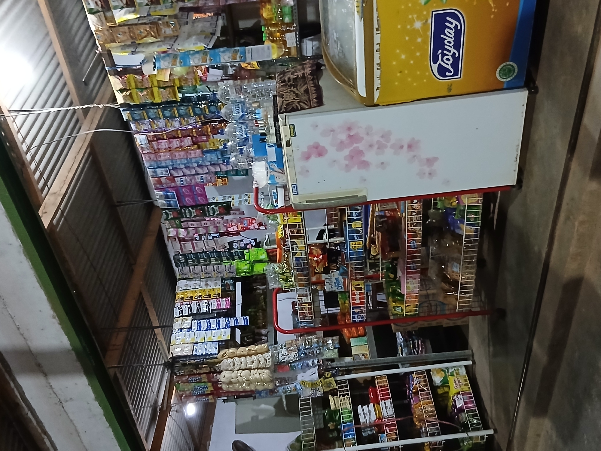 This image captures the interior of a small convenience store, likely a warung, in Setia Bakti, Indonesia. The scene is filled with various goods displayed on shelves and hanging from the ceiling. Rows of packaged snacks, drinks, and household items are visible, suggesting a well-stocked local shop. Two refrigerators are present, one white with a floral decal and another yellow with the "Joyday" logo, likely for chilled products. The overall ambiance is that of a typical Indonesian neighborhood store, serving the local community. The lighting appears to be a mix of overhead fluorescent lights and natural light filtering in. The floor is concrete and slightly worn, reflecting the everyday use of the space.