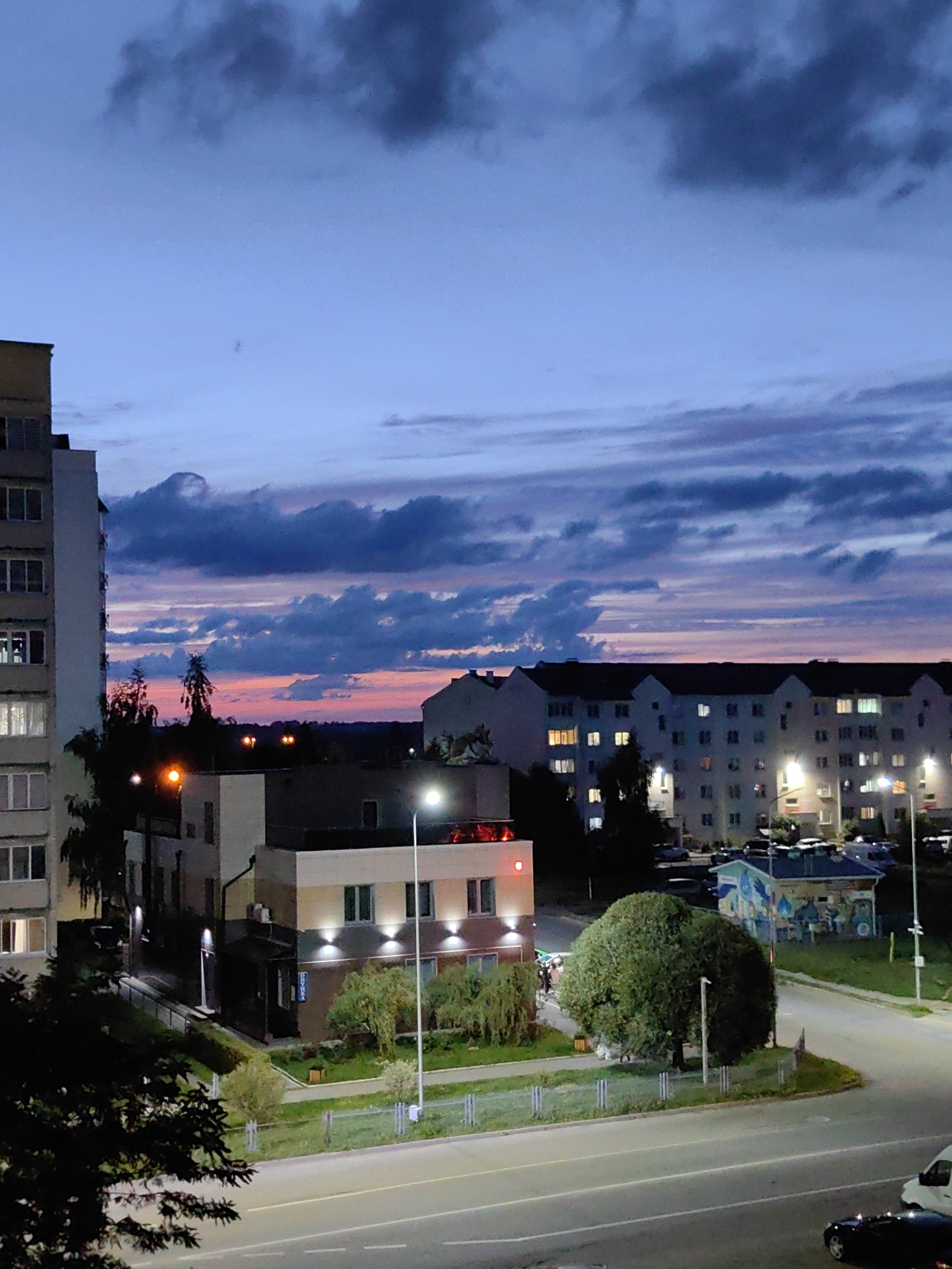 The image captures a twilight scene in Fanipal, Belarus, looking towards a residential area with multi-story buildings. The sky is a gradient of deep blue and purple, transitioning to a soft pink and orange horizon, indicative of sunset or sunrise. Dark, textured clouds streak across the upper portion of the sky.
In the foreground, a paved road with white lane markings is visible, with a portion of a white van and a black car on it. To the left, a tall, light-colored apartment building with visible windows is present. Adjacent to this, a low-rise building with a light-colored facade and windows, some illuminated from within, is situated. In front of this building, there are trees and greenery.
Further into the scene, a row of connected apartment buildings forms the mid-ground. These buildings have multiple floors and numerous illuminated windows. Streetlights, some with a warm orange glow and others with white light, are present, illuminating parts of the street and buildings. A large, rounded tree with dark foliage stands prominently in the mid-ground, casting a shadow. Behind the tree, a smaller building with a mural or graffiti art on its side is visible. A fence runs along the edge of a grassy area. The overall impression is a calm, urban residential environment at the transition between day and night.