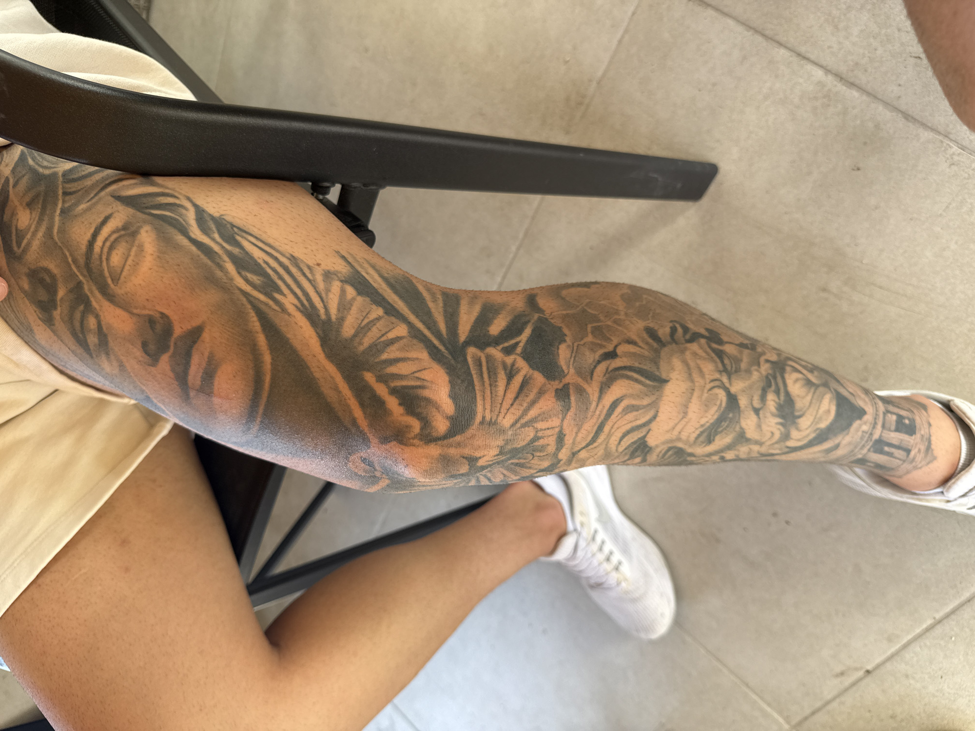 A person with a tattoo sleeve on their left leg is sitting on a chair indoors. The tattoo features a detailed black and grey design, including faces, flowers, and other decorative elements. The person is wearing a light-colored pair of shorts and white sneakers. The chair has a black armrest, which is partially obscuring the tattoo on the upper part of the leg. The floor is tiled in a light, neutral color. The location is unknown.