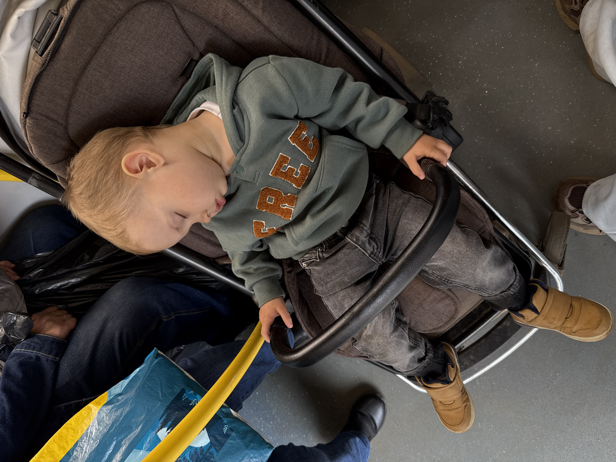 This overhead shot captures a young child soundly asleep in a dark brown stroller, likely within a public transport vehicle in Rusanovo, Russia. The child, with light blonde hair, is lying on their back with their head tilted slightly to the left, eyes closed, and mouth slightly open, indicating deep slumber. They are dressed in a green hooded sweatshirt featuring large, stitched brown letters, prominently displaying "BREA" across the chest, paired with dark grey jeans and tan-colored, high-top sneakers with velcro straps.
The stroller is dark brown with cushioned padding, set on a grey, speckled floor that suggests the interior of a bus, tram, or train. Around the stroller, elements of travel are visible. To the left, an adult's legs are seen, clad in dark blue jeans, with a black shoe peeking out. A black plastic bag and a blue shopping bag with a landscape graphic are also nestled beside the stroller. A yellow bar, possibly a handrail or safety strap, runs diagonally across the lower left of the frame. In the background, another person's legs in light-colored pants and brown sneakers are partially visible. The overall scene conveys a sense of peaceful transit, with the child comfortably resting during their journey.