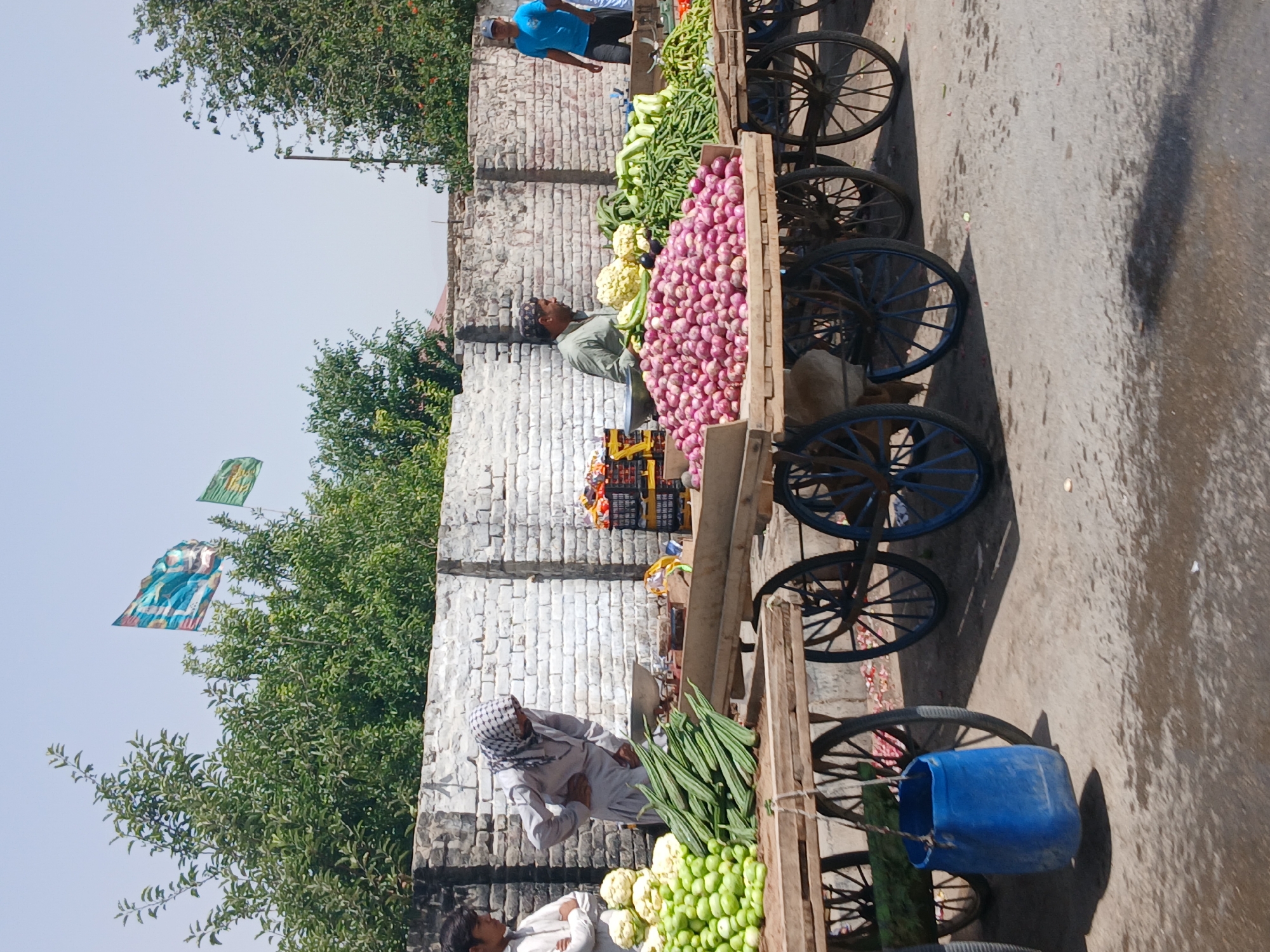 The image depicts a street scene in Quetta City Tehsil, Pakistan, showcasing a roadside vegetable market. Several vendors are present, standing near their wooden carts filled with produce. The carts are arranged in a row along a wall constructed of white brick. The vendors offer a variety of vegetables including onions, caulifower and long green squash. One vendor, wearing a headscarf, is seen interacting with a customer. Another vendor is seen in a blue shirt. Trees with green leaves and colorful flags are visible in the background. The ground appears to be asphalt. The light conditions suggest it is daytime.