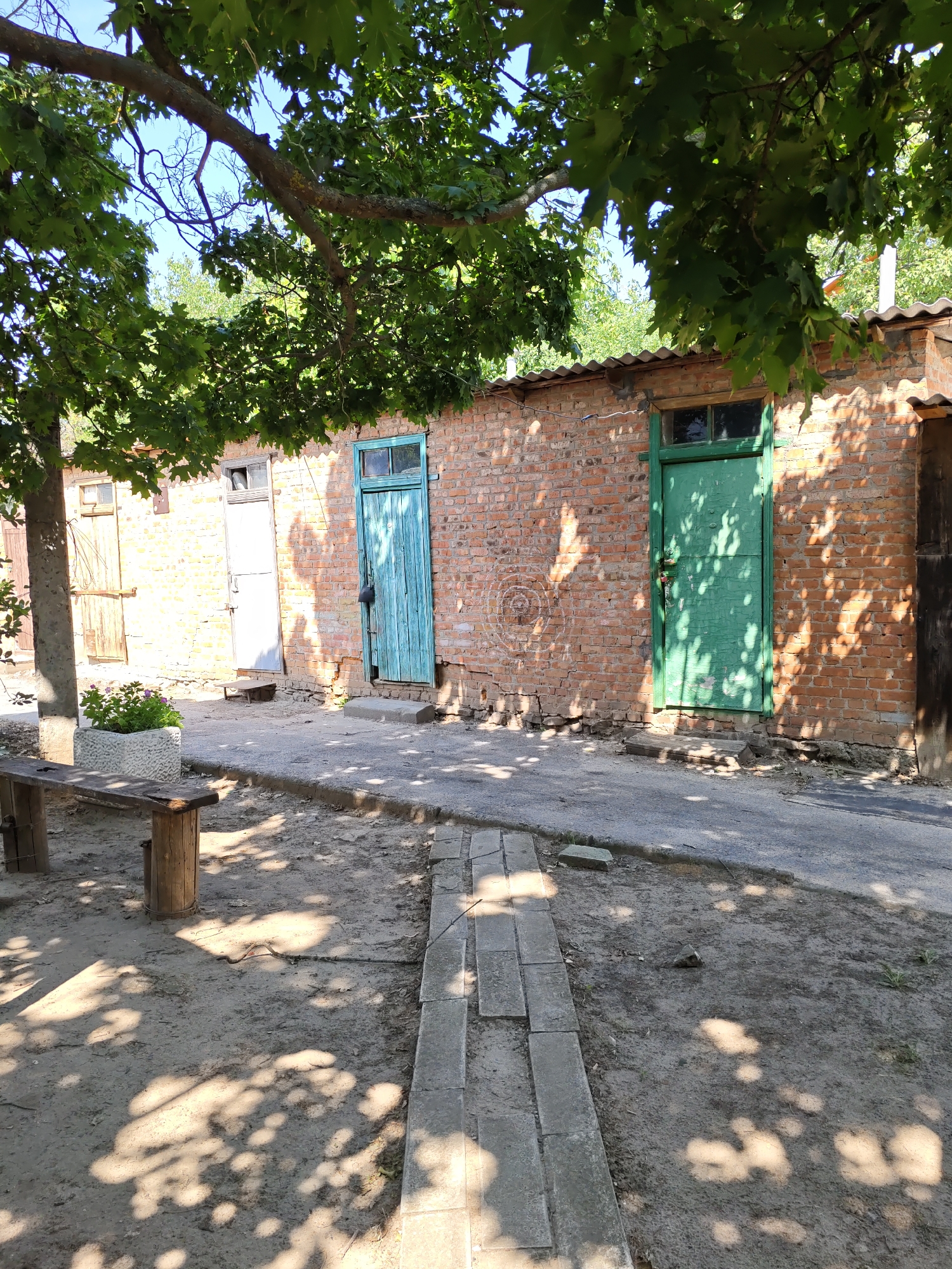 The image shows a view of a brick building with two green doors and two wooden doors. A tree branch with green leaves hangs over the scene from the top. In front of the building, there is a path made of concrete slabs. To the left of the path is a wooden bench next to a planter box with green plants. The ground is covered with dirt and scattered leaves. The scene is in Pukhlyakovskiy, Russia.