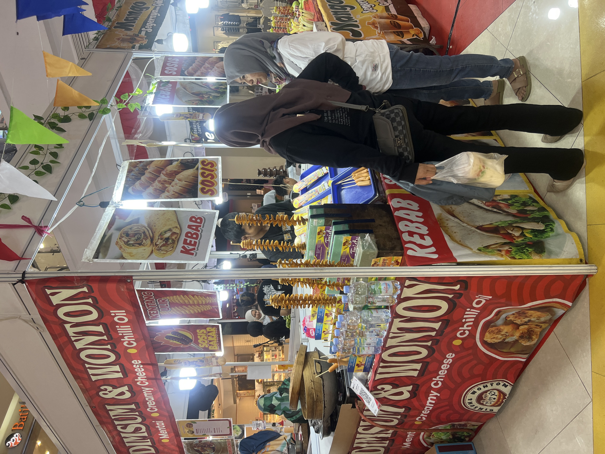 The image shows a food stall at a market in Banyurojo, Indonesia. The stall is selling dim sum and wonton, as indicated by the large red banner at the front. There are several types of food displayed, including kebabs, sausages, and what appears to be fried potato spirals. Bottles of water are lined up on the counter. Two women, wearing headscarves, are standing in front of the stall, with one of them holding a plastic bag of food. Another person, likely a vendor, is visible behind the counter. The stall is decorated with colorful flags and leafy green vines. In the background, other stalls and shoppers can be seen, suggesting a busy market environment.