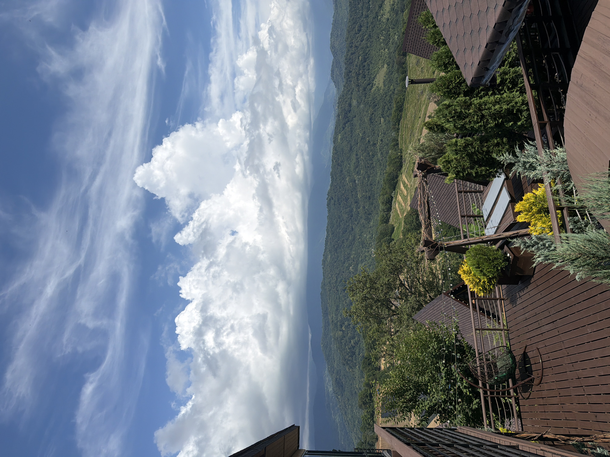 A panoramic view from a wooden deck overlooks a vast, tree-covered mountain range under a dynamic sky with scattered clouds. The deck, made of dark brown planks, is furnished with a woven hanging chair, planters with yellow and green shrubs, and a swing-like structure adorned with string lights. In the foreground, the edge of a round wooden table is visible. Several houses with dark brown tiled roofs are scattered across the landscape. The distant mountains are layered, with the closest covered in dense green forest and further ranges receding into hazy blue. The sky exhibits both bright white cumulus clouds and wispy cirrus formations against a clear blue backdrop. The location is identified as Nizhnyaya Shilovka, Russia. There are no people or animals visible in the image.