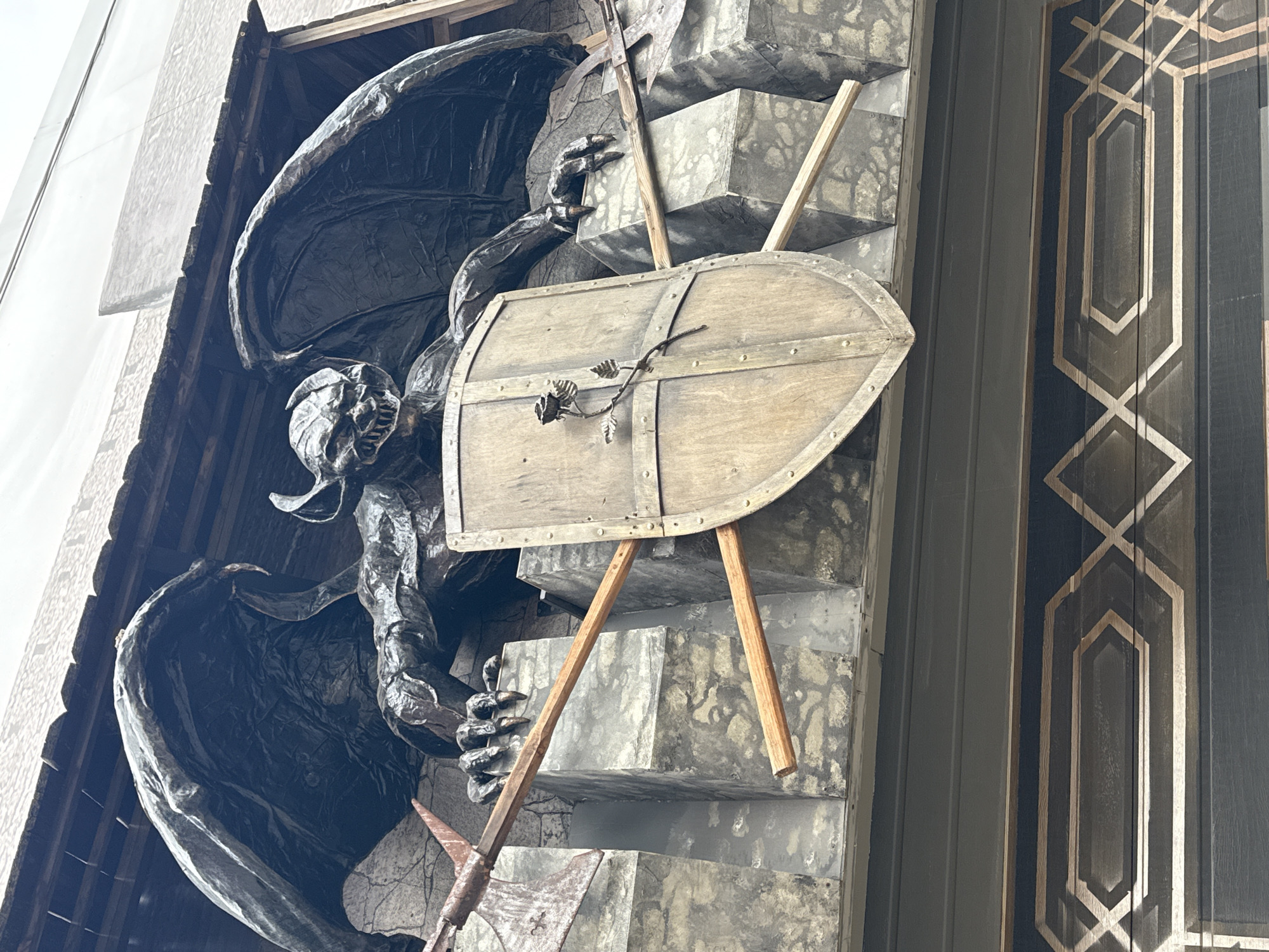 The image shows a close-up of a dark, weathered gargoyle sculpture positioned on the side of a building. The gargoyle has large, leathery wings and sharp claws. It is holding a wooden shield with metal rivets, decorated with a metal rose and vine. An axe is also visible, leaning against the shield. The background appears to be a concrete or stone wall, possibly part of the building's facade. To the right of the sculpture, a section of the building features a decorative panel with a geometric pattern of light-colored lines against a dark background. The overall impression is of an architectural detail with a fantastical or gothic theme. The lighting suggests it is daytime, with natural light illuminating the scene. The gargoyle's expression is difficult to discern due to its stylized nature, but it evokes a sense of strength and possibly menace. The location context is Perm, Russia.