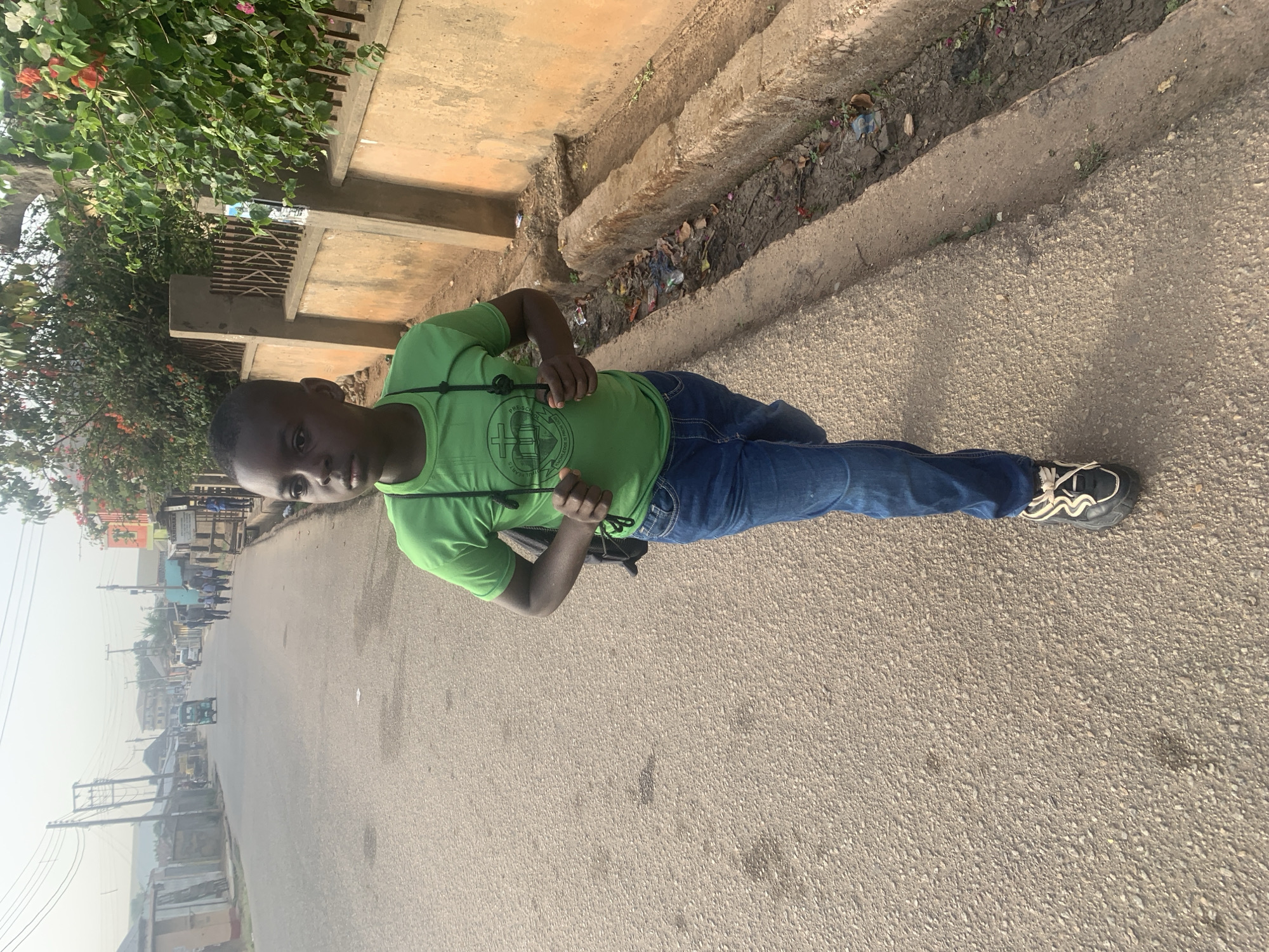 A young person stands on a paved street in Jos, Nigeria, looking directly at the camera. The individual wears a bright green short-sleeved t-shirt, blue jeans, and dark athletic shoes with white detailing. A black strap, possibly from a bag or camera, crosses their shoulder.
The street surface is light grey asphalt. To the right, a concrete curb delineates a dirt gutter containing some litter and debris. To the left, lush green foliage with hints of red blossoms grows behind a concrete wall or fence. In the background, the street extends into a hazy distance lined with buildings, power poles, and distant figures. The sky is bright but appears overcast or hazy.
The person is posing for a photograph. The scene is illuminated by daylight, suggesting mid-morning or afternoon. On the green t-shirt, a circular logo is visible, featuring a cross symbol above an open book. Around the circumference of the logo, text can be partially discerned, reading "PRESENTS" at the top, "FOR KNOWLEDGE AND UNDERSTANDING" in the middle, and "TRUE HEARTS" at the bottom.