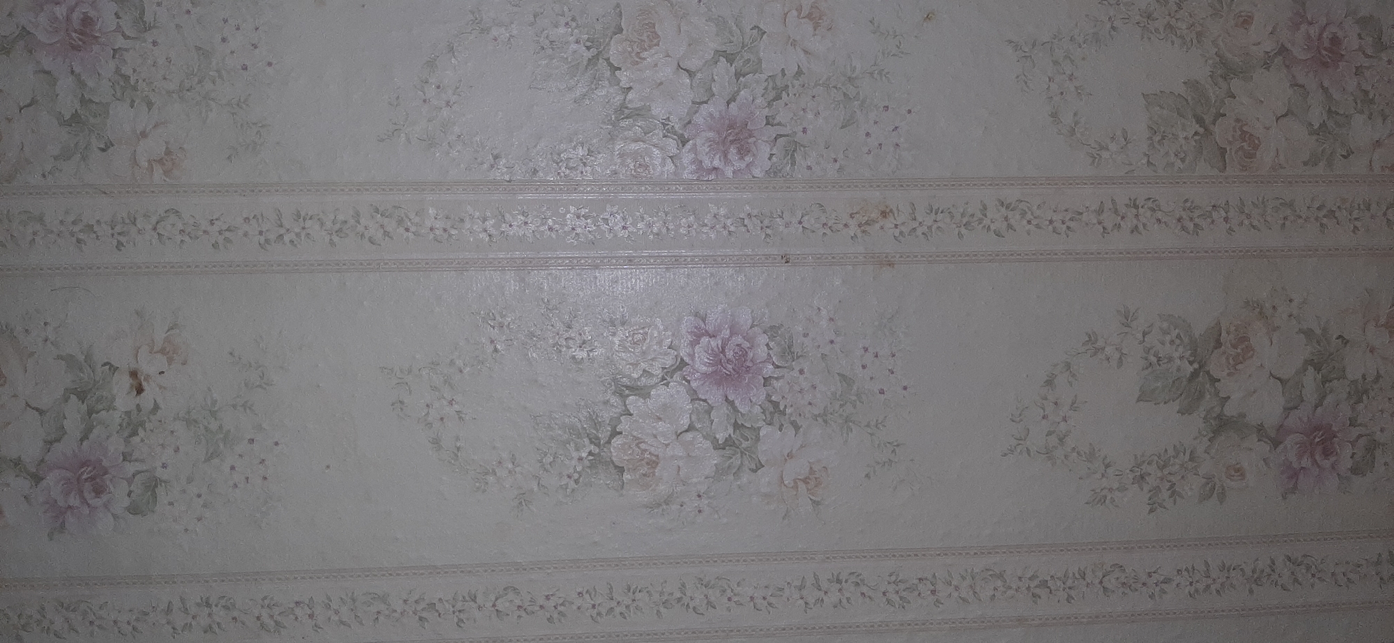 The image displays a close-up view of floral wallpaper with a pale, off-white background. Vertical striped patterns, featuring a delicate chain of small flowers and leaves, run down the wallpaper. Interspersed between these stripes are larger, faded floral motifs, primarily depicting peonies and other rose-like blooms in shades of pink, white, and pale green. The wallpaper appears aged, with some subtle discoloration and minor imperfections visible. The scene is consistent with interior wall covering. The location of Yessentukskaya, Russia, is mentioned but not visually represented in the provided image.