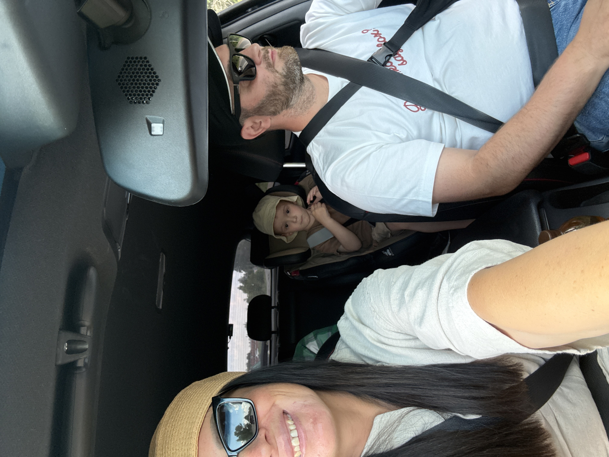 This is a selfie taken inside a car. In the foreground, on the left, a woman with long dark hair, wearing a straw hat and sunglasses, smiles at the camera. Her left arm is visible, showing tanned skin with freckles. In the middle of the frame, a young boy wearing a tan hat sits in a car seat. To the right, a man wearing a black baseball cap and sunglasses drives the car. He has a beard and is wearing a white t-shirt with red lettering and blue jeans. The car's interior is visible, including the dashboard, rearview mirror, and seatbelts. The background shows a glimpse of trees and a road, suggesting the car is in motion. The image does not contain any information that would allow for a specific location within the city of Chaikovsky, Russia.