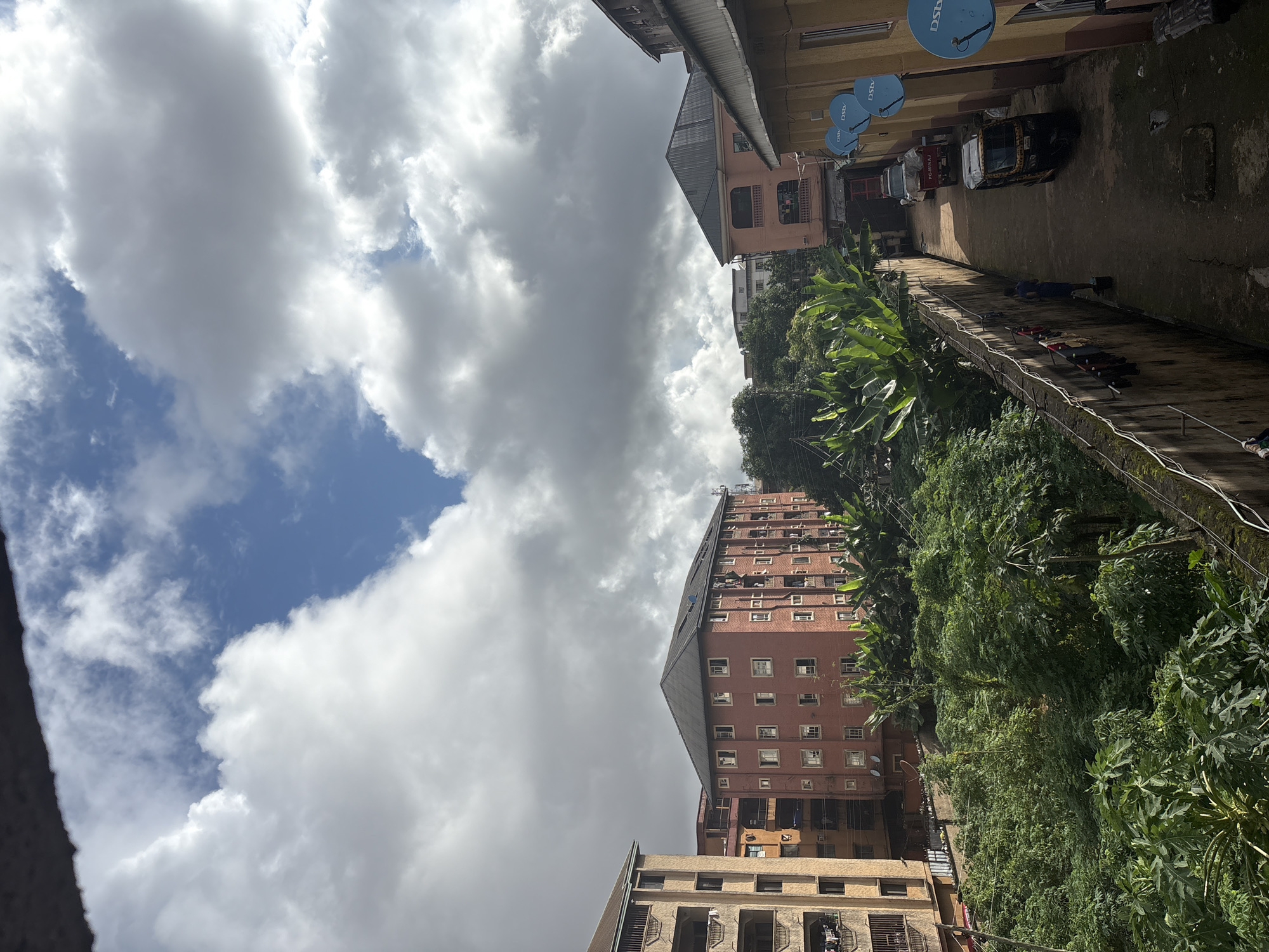 This image depicts a view of Onitsha, Nigeria, characterized by a mix of urban structures and lush vegetation. The sky is filled with dramatic, puffy white and gray clouds against a bright blue backdrop. In the mid-ground, several multi-story buildings are visible, including a prominent reddish-brown brick apartment building with numerous windows and balconies. To the left, another apartment building, lighter in color and with balconies, is also present. A dense layer of vibrant green foliage, including large-leafed plants, occupies the foreground and extends between the buildings, suggesting a tropical climate. A narrow walkway or alleyway is visible on the right, bordered by a concrete wall with clothes hanging on a line. Along this walkway, a yellow and black auto-rickshaw (Keke Napep) is parked, and further down, a white truck. A single person, wearing a blue shirt, stands in the alleyway near the wall. Several satellite dishes, identified by the "DSTV" logo, are mounted on the side of a yellow building on the right. The overall impression is one of a densely populated urban environment with significant green spaces.