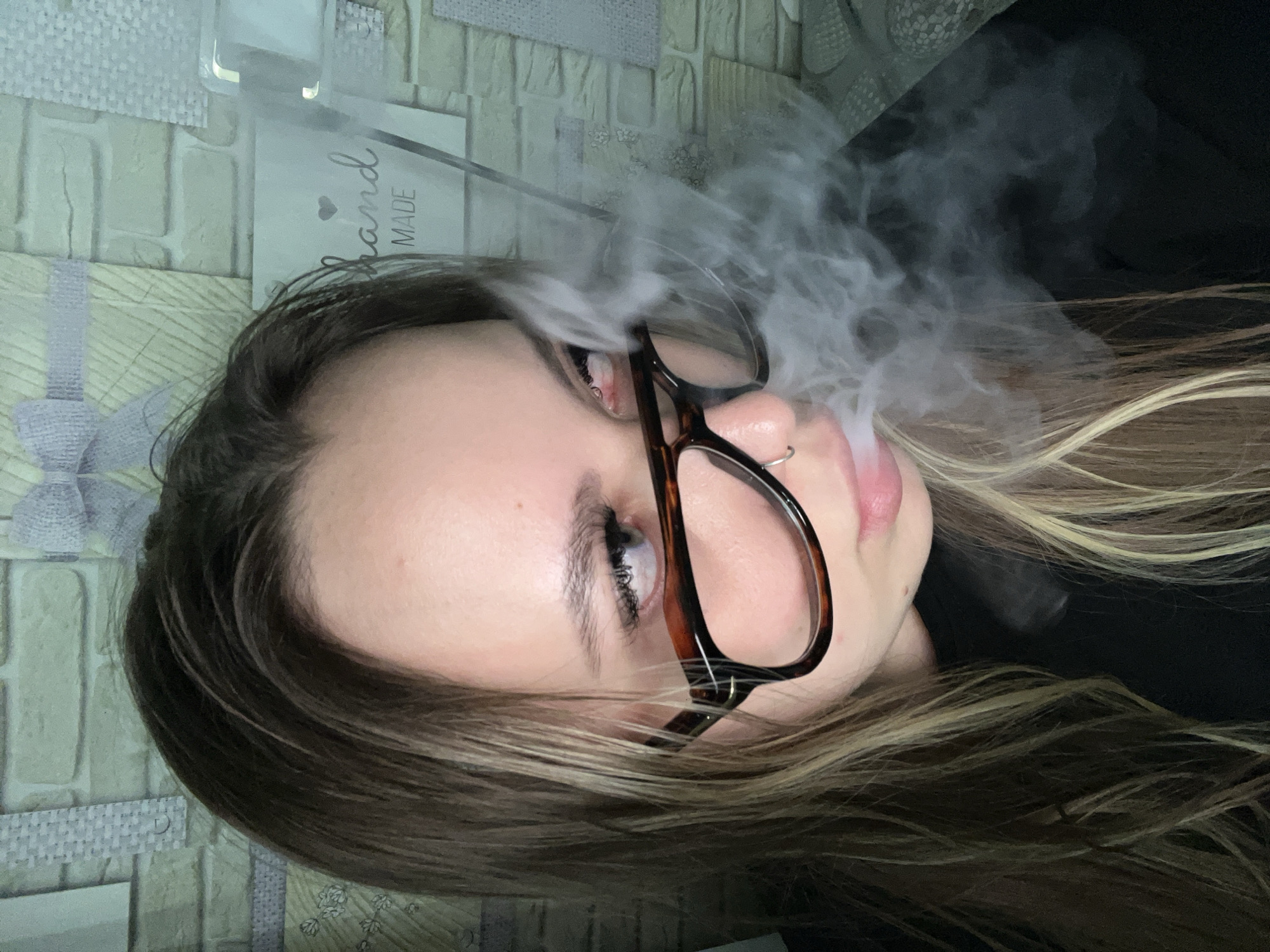 A woman with brown hair highlighted with blonde streaks, wearing tortoiseshell-framed glasses and a small nose ring, is positioned in front of a decorative brick-patterned wall. She is partially obscured by a plume of smoke emanating from her mouth. The setting is an indoor location, identifiable as Port-Katon, Russia.