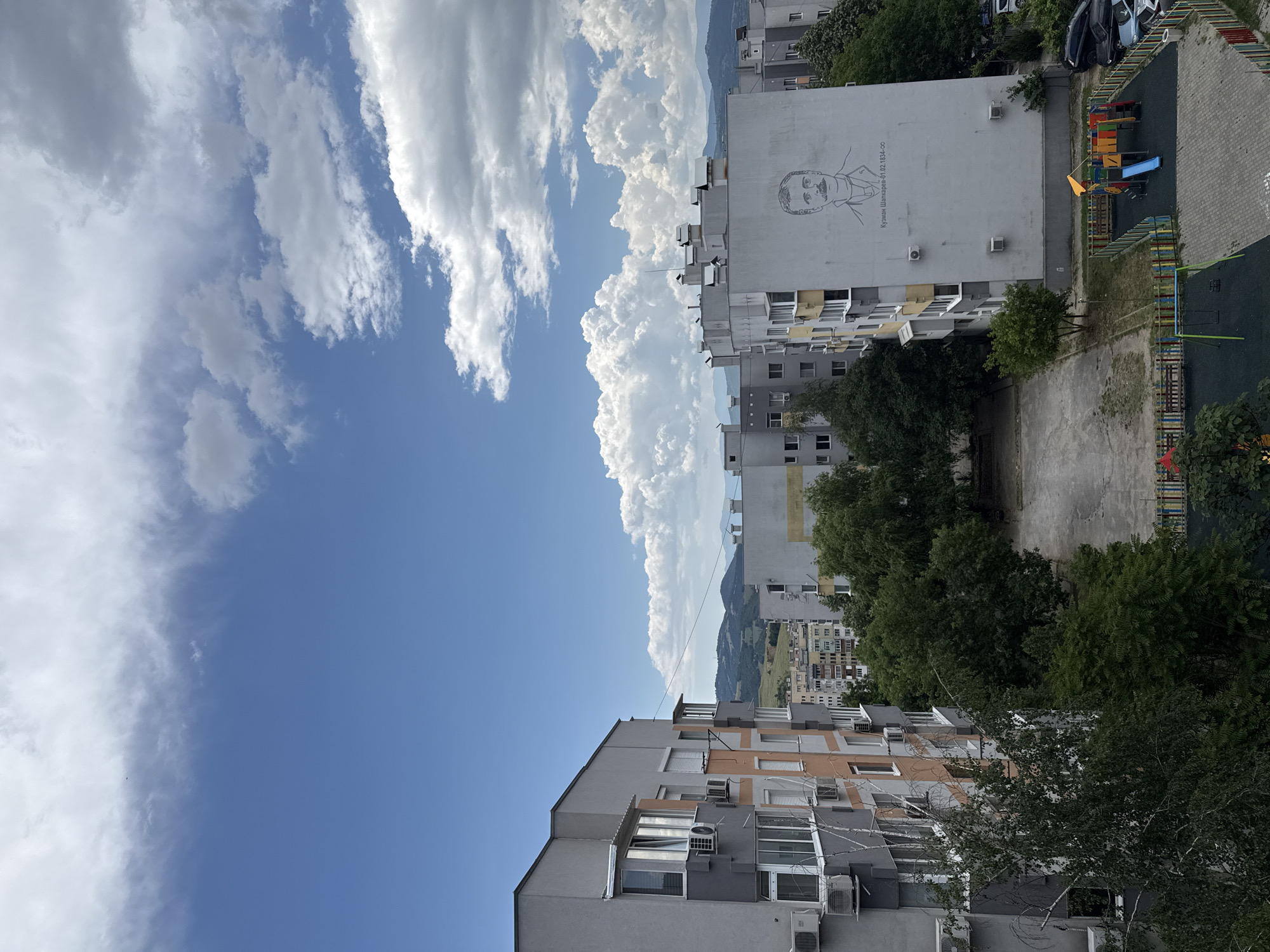 This image presents an elevated view of a residential area in the city of Blagoevgrad, Bulgaria, under a partly cloudy sky. The scene is dominated by multi-story residential buildings, primarily constructed in a utilitarian socialist-era style, characterized by their grey facades. Some buildings feature sections painted in peach-orange or pale yellow, and numerous external air conditioning units are affixed to their walls, alongside various windows and balconies.
In the foreground to the left, a tall grey building with peach-colored accents is prominent, with a slender tree having light-colored bark in front of it. Towards the right, a distinctive grey building showcases a large, black-and-white mural of a man with a mustache and cravat, identified by the Bulgarian text below as "Кузман Шапкарев - 01.02.1834-ос" (Kuzman Shapkarev - 01.02.1834-born). This building also has multiple air conditioning units. Other similar residential blocks are visible receding into the middle ground.
At ground level, an unpaved, irregular dirt or gravel area is visible towards the center-right. Adjacent to it, a colorful children's playground is enclosed by a multi-colored fence, featuring various play structures including slides, swings, and climbing elements, set on a dark, soft ground surface and paving stones. To the far right, a small parking area holds several parked cars, including a black SUV, a blue sedan, and a white vehicle. Lush green trees and bushes are interspersed throughout the urban landscape, particularly around the ground-level features and between buildings.
In the distance, rolling green hills and mountains define the horizon. The sky overhead transitions from clear blue with wispy clouds at the upper left to areas with more prominent, fluffy cumulus clouds on the right. No people are visible in the scene.