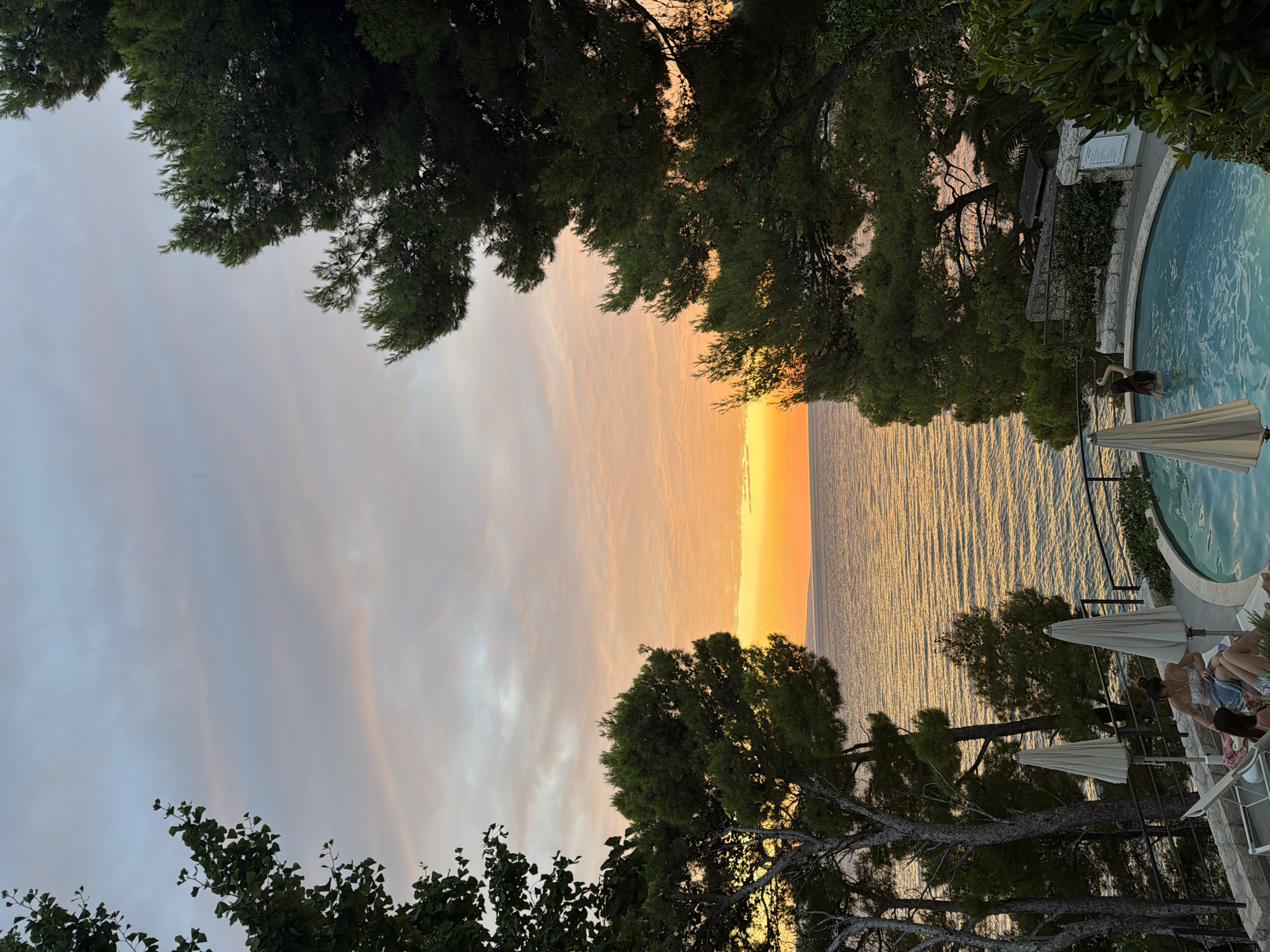 The image depicts a sunset over the Adriatic Sea, viewed from a terrace in Općina Brela, Croatia. The sky is a gradient of pale blue and soft orange hues, with wispy clouds. Two people are visible near a circular swimming pool filled with clear blue water. One person, wearing a floral top and denim shorts, is sitting on a lounge chair, looking down. Another person, with long dark hair, is partially submerged in the pool, with their back to the camera, appearing to wave an arm. Two large, cream-colored beach umbrellas stand near the lounge chairs. Lush green pine trees frame the scene, with their branches reaching towards the sky. The sea stretches out to the horizon, reflecting the warm colors of the sunset. A stone wall and some greenery form the edge of the terrace.
