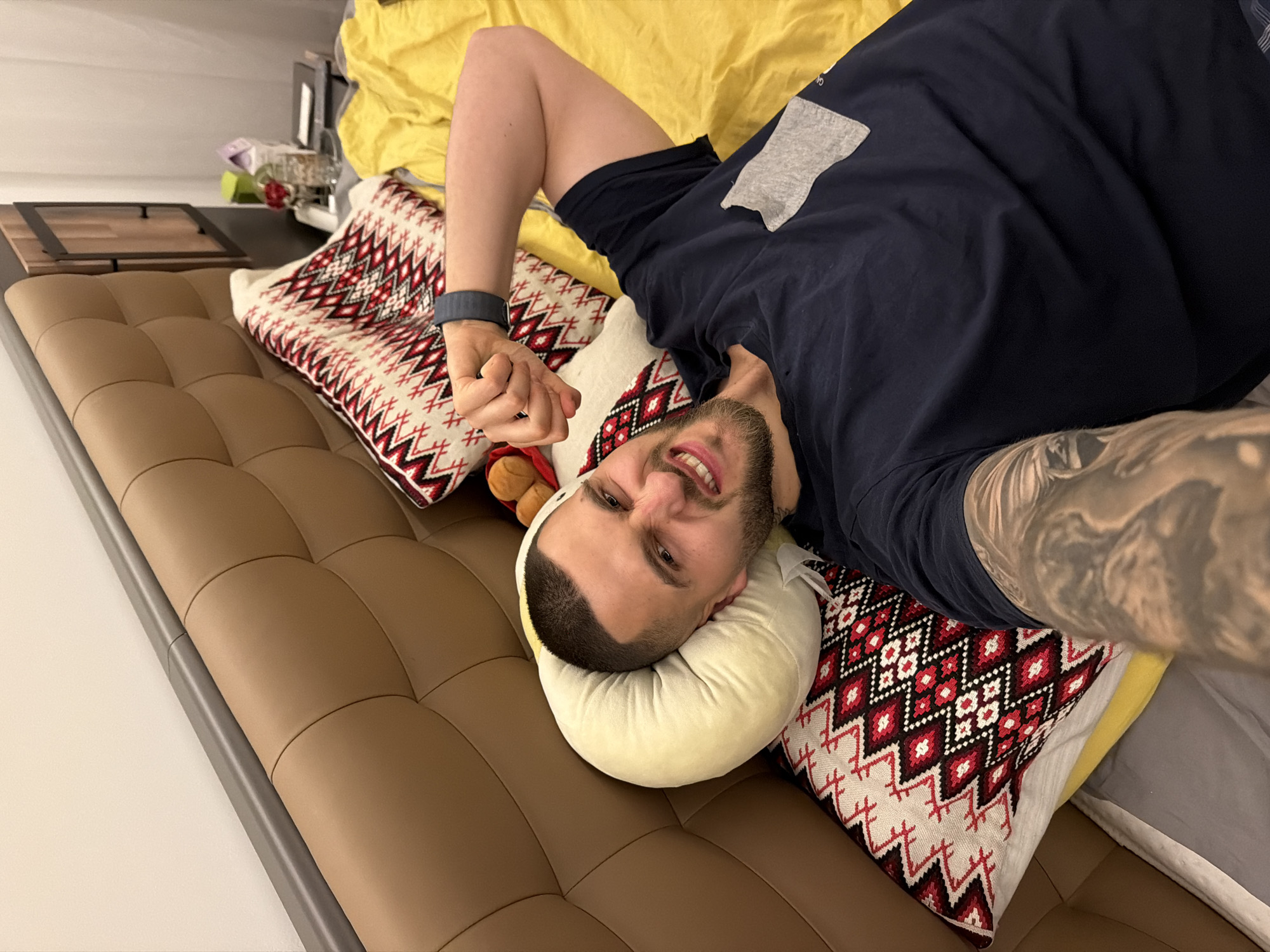 A man with a buzzcut, light complexion, and a short beard is lying on his back on a brown leather couch. He is wearing a dark blue t-shirt with a gray patch on the shoulder. His head rests on a yellow pillow, and he is surrounded by decorative pillows with red and white patterns. A grey smartwatch is visible on his left wrist, and a tattoo is visible on his right arm. The background includes a yellow blanket and other items on a shelf. The location is likely an interior setting, possibly a residence, but there is no evidence to suggest it is located in Sofia, Bulgaria.