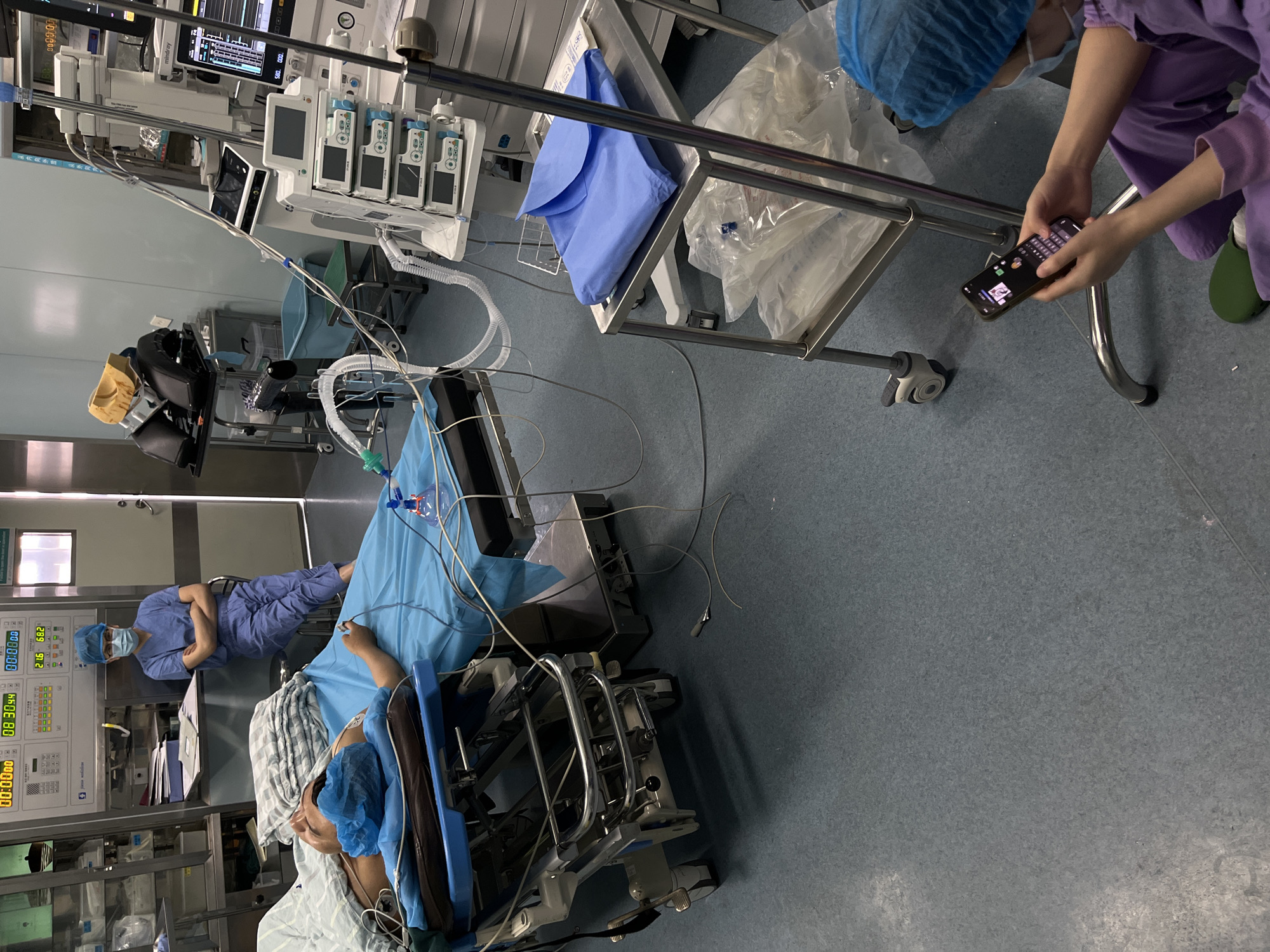This media file captures an indoor clinical setting, likely an operating room preparation or recovery area within a hospital in Yuzhong District, China. The scene is brightly lit by artificial light.
In the foreground and center, a patient lies on a medical gurney, covered by a blue surgical sheet and a striped blanket, wearing a blue surgical cap. Wires and tubes connect the patient to various medical equipment. To the right, a medical professional in purple scrubs, a blue surgical cap, and green clogs is crouching, intently looking at and interacting with a smartphone held horizontally. On the left side of the frame, another medical professional, wearing blue scrubs, a blue surgical cap, and a mask, stands with arms crossed, leaning against a white medical console, appearing to observe or wait.
The room is filled with medical equipment. Several monitors, one branded "Mindray," display digital readouts including waveforms, "00:00:00" timers, and numerical data such as "08 30.94," "20.9," and "21.6." A rack with four infusion pumps is visible, along with tubes, hoses, and IV stands extending towards the patient. A small metal cart holds blue drapes, and another larger wheeled cart with clear plastic bags is near the crouching professional. The floor is gray, and the walls are light-colored. The overall atmosphere appears calm and routine, with no immediate signs of distress or urgency.