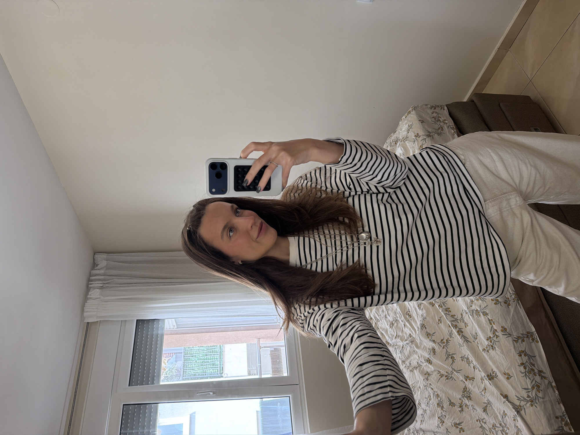 A woman is taking a selfie with a cell phone in a room in Budva, Montenegro. She is wearing a striped black and white shirt and white pants. She is standing in front of a bed covered with a floral patterned bedspread. A window is behind her, showing a building outside.