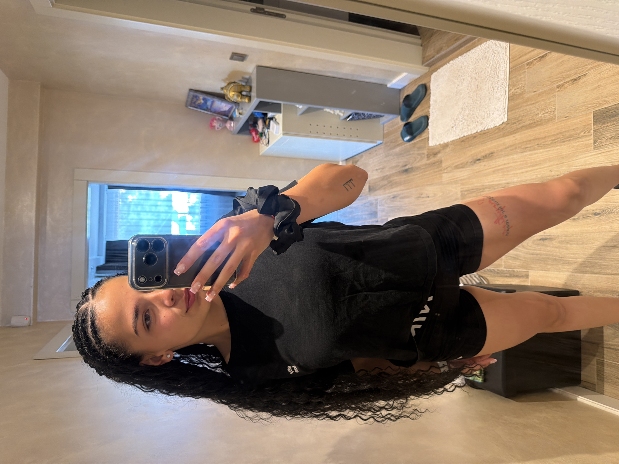 A young woman with long, braided dark hair and an athletic build is taking a mirror selfie in what appears to be a hallway or entryway. She is wearing a black t-shirt and black shorts. Her right arm is raised, holding a smartphone. On her right forearm, there is a tattoo. To her left, a black storage bench is partially visible. In the background, a doorway leads to another room, and a shelving unit holds decorative items. The floor is made of light-colored wood planks, and a small white rug is positioned in front of the shelving unit. The walls have a textured, light beige finish. The scene is well-lit, possibly by natural light from the adjacent room. There are no visible indicators that this location is in Gabrovo, Bulgaria.