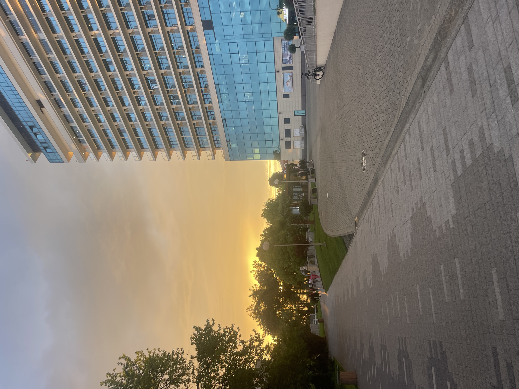 The image shows a street scene in Sochi, Russia, during sunset. On the left, a tall, modern building with multiple floors of balconies and a blue glass facade reflects the golden sky. A pedestrian walkway with gray cobblestone paving runs along the right side of the image. Trees with green foliage line a park area between the walkway and the sunset. Several people are walking in the park. A bicycle is parked next to the building on the right. The overall atmosphere is warm, bathed in the golden light of the setting sun.