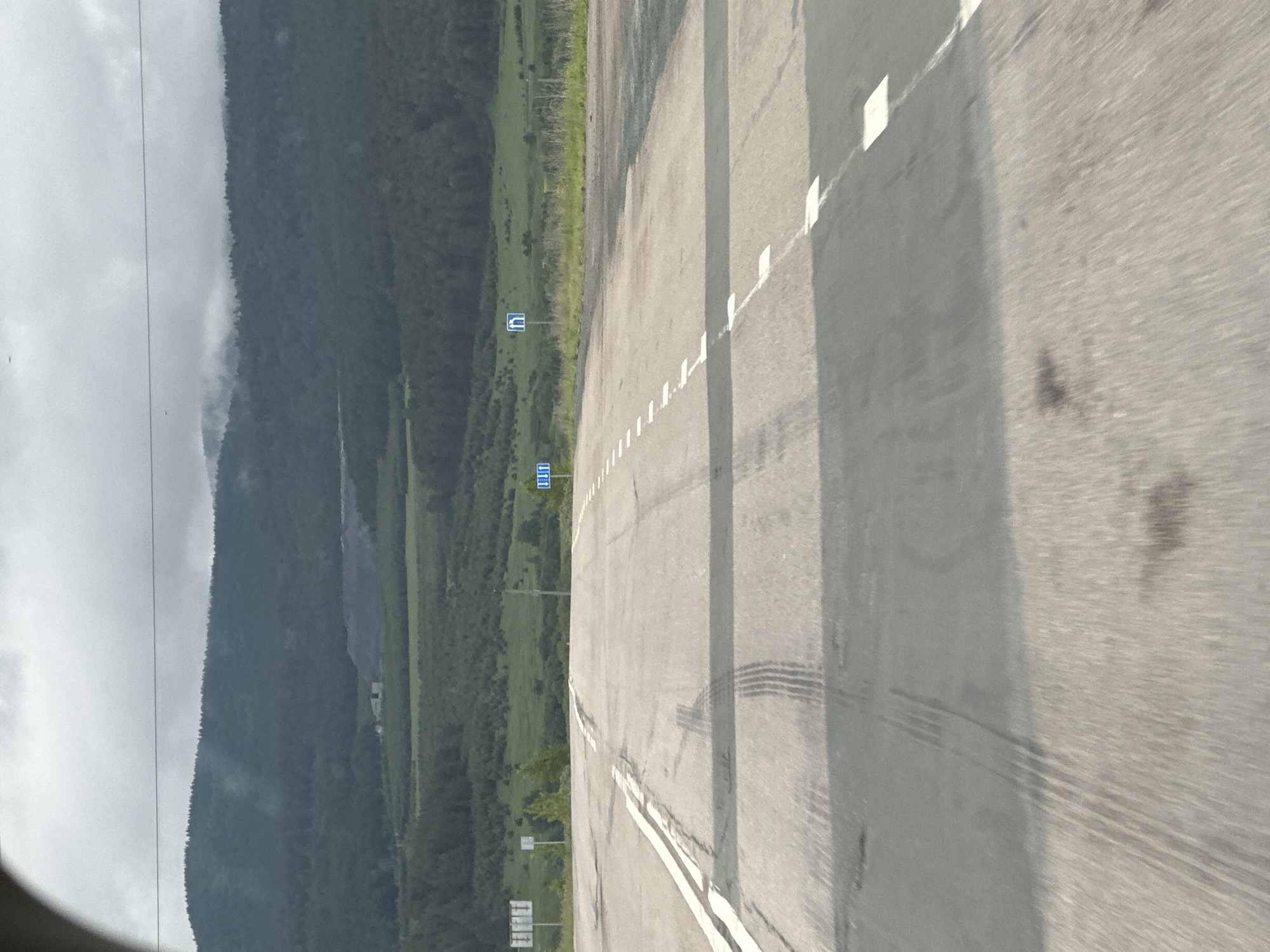 The image shows a road perspective with a cloudy sky and rolling hills in the background. The road has white lane markings and tire tracks are visible on the asphalt. In the distance, there are trees and some structures. There are also road signs, one of which appears to be a blue sign with a white symbol indicating a turn or intersection. The scene suggests an outdoor, rural setting. It is not possible to determine the specific location within Lomovka, Russia from the provided image.