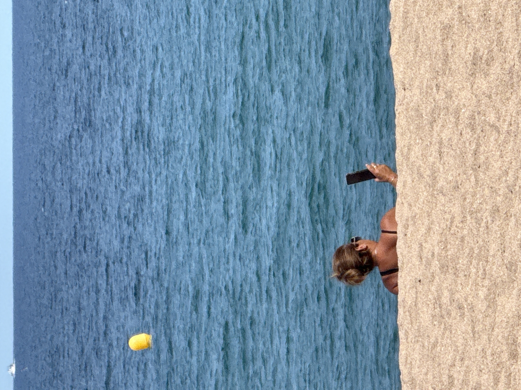 A woman with blonde hair pulled up in a bun sits on a sandy beach, facing away from the camera and towards the blue sea. She is wearing sunglasses and a dark bikini top. In her right hand, she holds a black smartphone, seemingly taking a picture or video. The water is calm with gentle ripples, and a yellow buoy is visible in the distance. The horizon line is clear, with a faint boat visible on the far left. The foreground is dominated by the textured sand of the beach. The scene appears to be a typical day at the beach in Blanes, Spain.