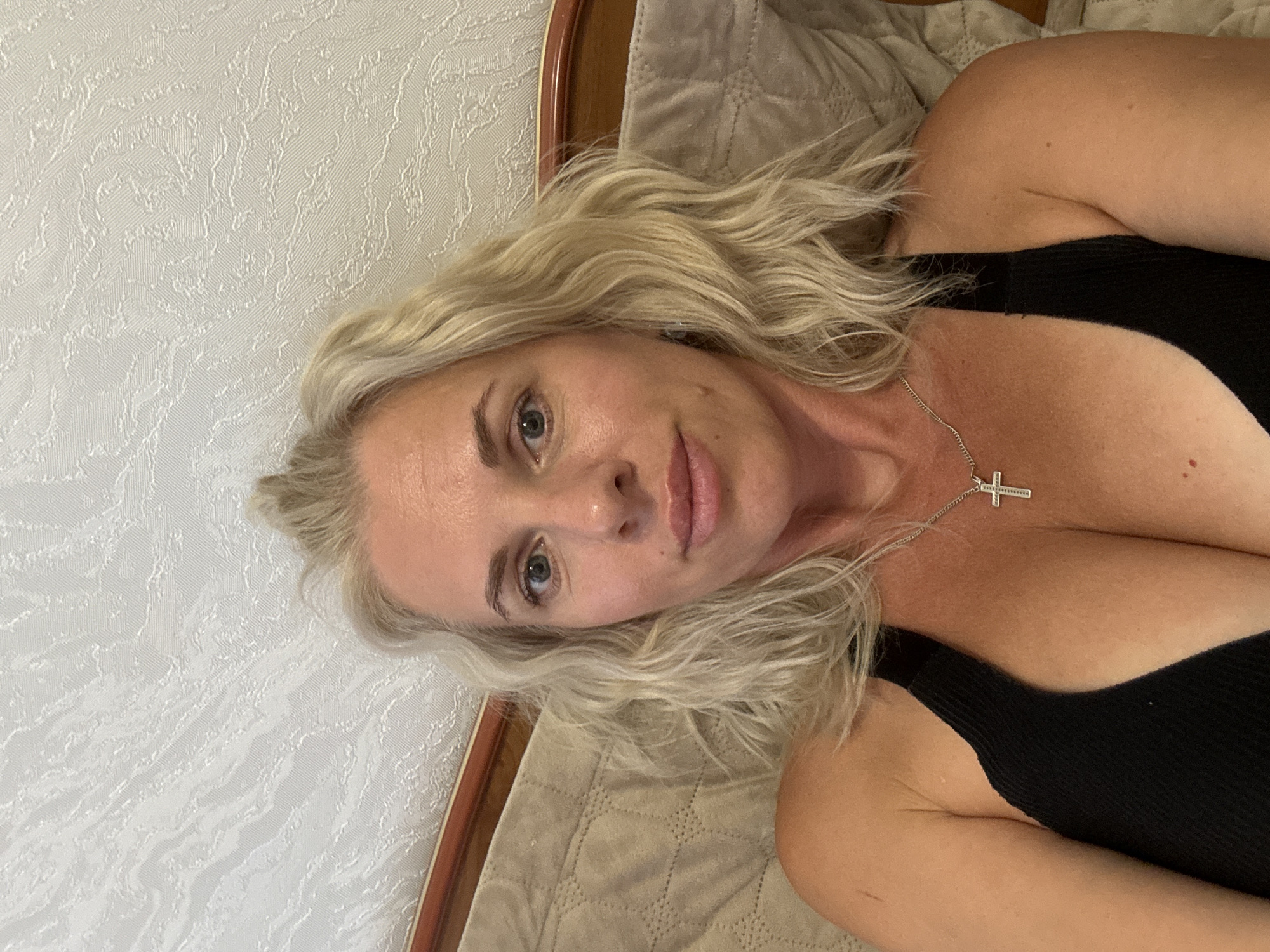 A woman with blonde hair is lying down and looking at the camera. She is wearing a black top and a silver cross necklace. The background is a textured wall and a bed. The image appears to be taken indoors, possibly in a bedroom.