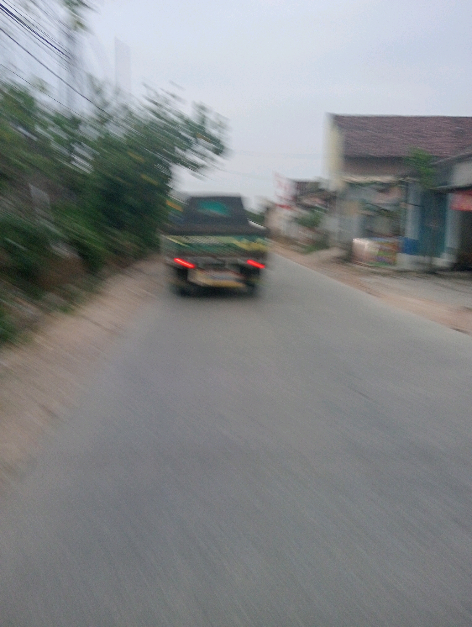 The image depicts a street scene in Pringsewu, Indonesia, with motion blur indicating movement. A pickup truck, possibly green and black, is visible on the right side of the road, its taillights illuminated. The truck appears to be moving away from the viewer. The road is paved and grey. On the left side of the road, there are trees and some structures, including what appears to be a building with a reddish-brown tiled roof. The sky is overcast and light-colored. The overall impression is that of a moving vehicle on a road in a somewhat rural or suburban area of Pringsewu.