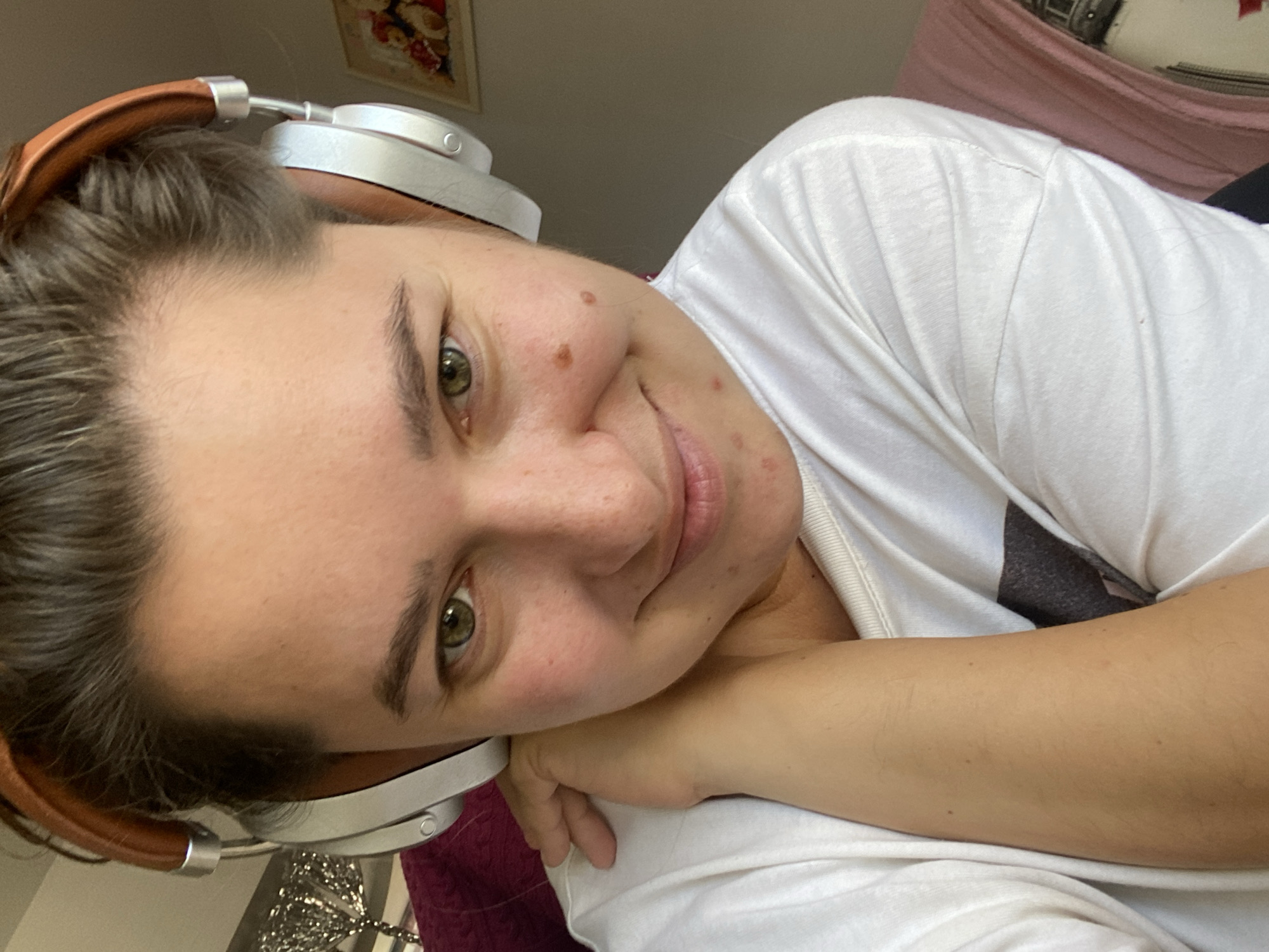 A close-up, overhead selfie of a young woman wearing silver and brown headphones. She is smiling and looking at the camera, with her left arm resting on her neck. She is wearing a white t-shirt. The background is blurred, but appears to be an indoor setting with a framed picture on the wall. There is no information about the location in the city of Perm, Russia.