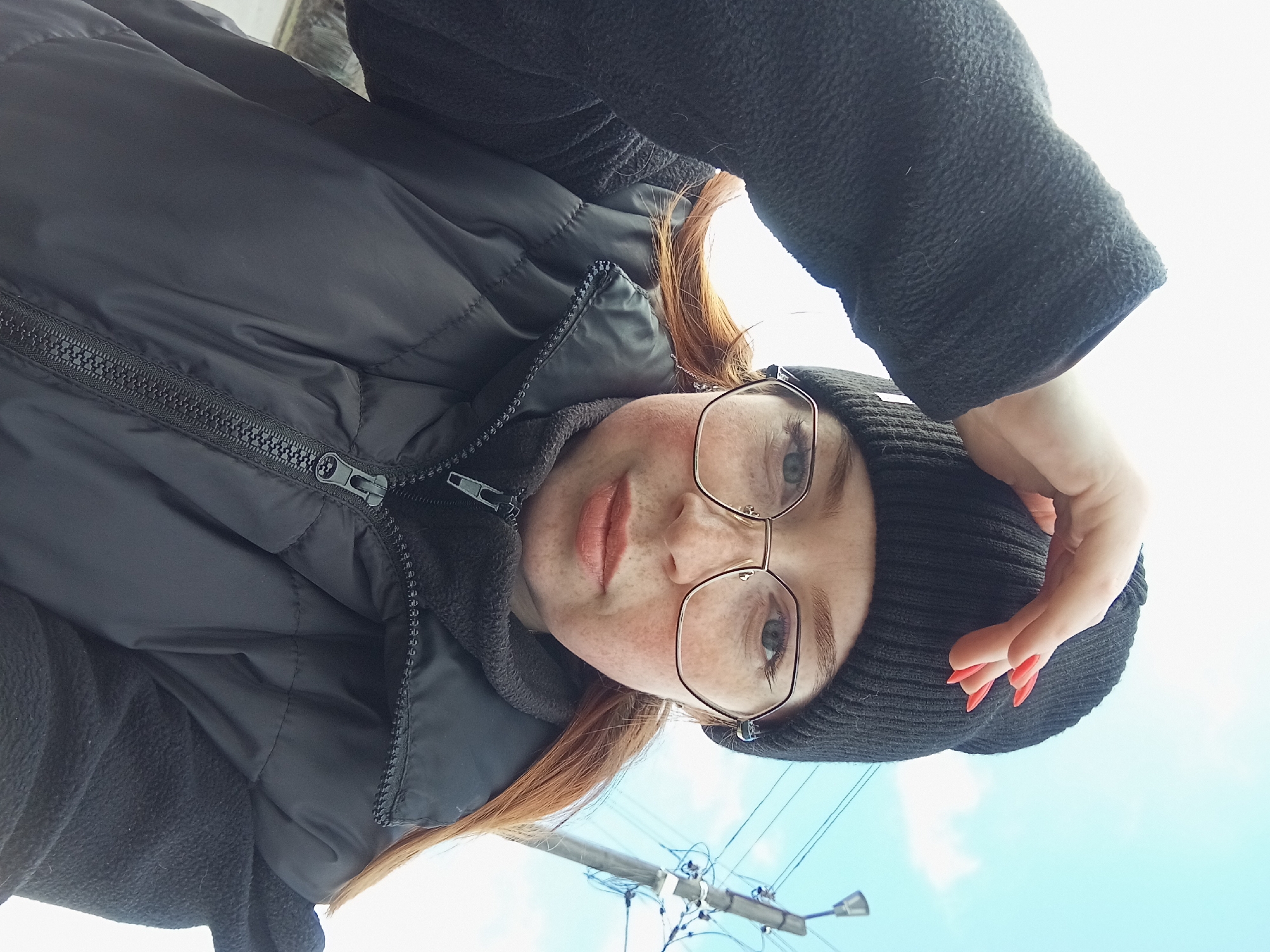 A low-angle outdoor selfie captures a young woman looking directly at the camera against a bright, light blue sky, possibly in Yurgamysh, Russia. She has fair skin, prominent freckles across her nose and cheeks, and reddish-brown hair peeking out from under a black knitted beanie. Her light-colored eyes are framed by round, gold-rimmed glasses. Her lips are slightly parted, revealing a light, peachy-pink hue.
She is dressed in a black padded or puffer jacket, with the main zipper clearly visible and pulled up. Underneath, a black fleece garment with a distinct textured fabric is visible, including its sleeve on the right side of the frame. Her left hand is raised towards her head, with fingers curled and fingernails painted a vibrant red.
The background primarily consists of a clear or slightly hazy bright blue sky. In the lower left portion of the image, a utility pole with various wires and fixtures is visible, suggesting an urban or suburban outdoor setting during daylight hours. The overall impression is a casual, personal photo taken on a bright day.