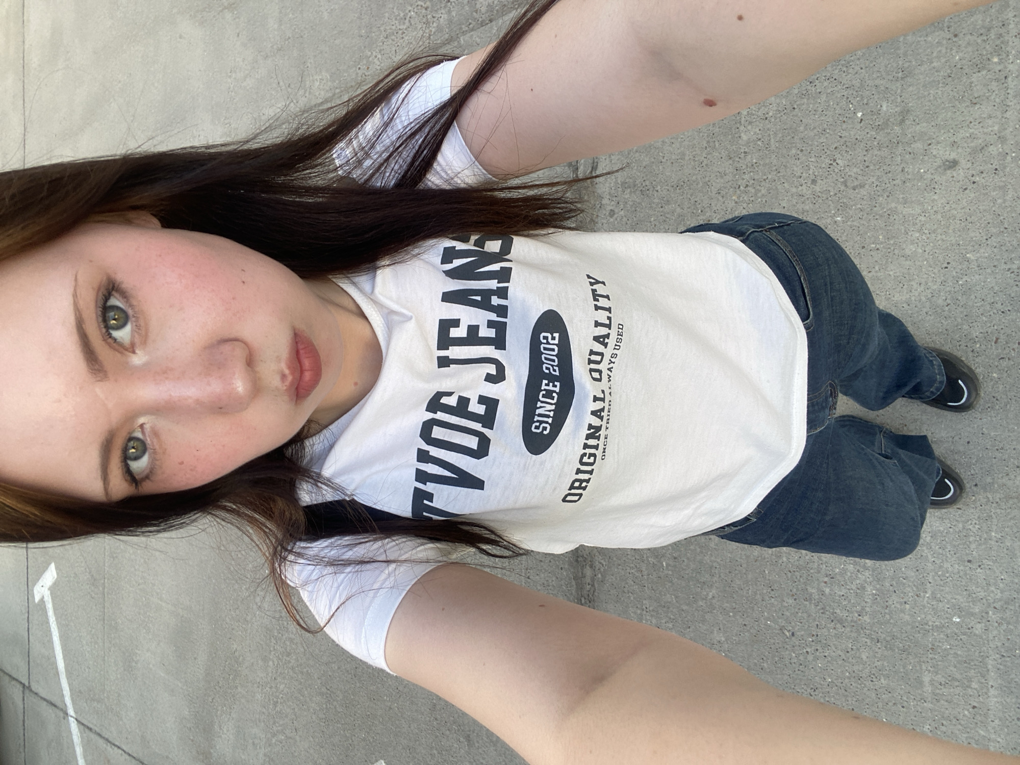A young woman is taking a selfie in an outdoor urban location. She is wearing a white t-shirt with "TVOE JEANS SINCE 2002 ORIGINAL QUALITY" printed on it, along with blue jeans and dark shoes. The location appears to be a concrete surface, possibly a street or parking area. The woman's hair is dark brown, and she has light skin with freckles.