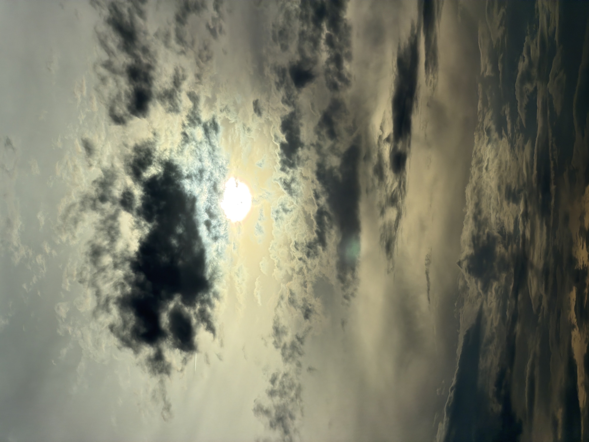 The image shows a cloudy sky with the sun partially obscured by dark clouds. The sun is a bright, white orb in the upper-middle section of the frame, surrounded by a hazy glow. The clouds are varied in texture and density, with some appearing dark and stormy, while others are lighter and more diffused. There are no people, subjects, objects, or actions visible in the image. The environment is solely the sky. The location is stated to be in Mramor, Bulgaria, but this cannot be visually verified from the image itself.