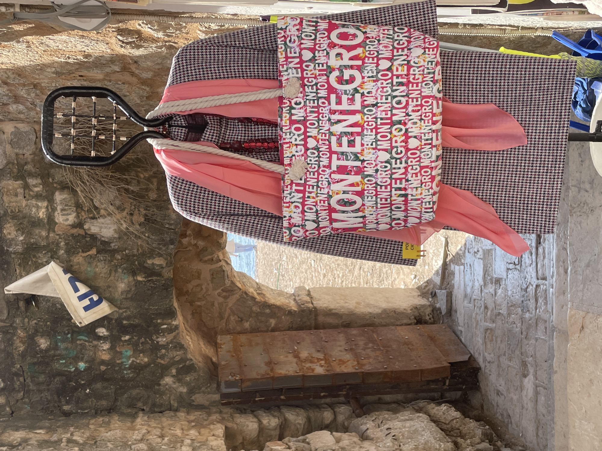 The image displays an outdoor scene in the Old Town of Budva, Montenegro, specifically within a stone-built passage leading towards a beach. In the immediate foreground, a mannequin torso or display stand presents a dark and white houndstooth patterned jacket. Around its neck, a bright coral-pink scarf is draped. Over the jacket, a large tote bag is displayed, featuring a repetitive pattern of "MONTENEGRO" in white capital letters and small heart icons on a red floral background, with thick white rope handles. Above the jacket, a dark, open-grid, racket-shaped metal structure serves as part of the display.
To the left of the display, a weathered stone archway forms a passage. A large, heavily rusted, rectangular metal door, approximately 2 meters tall, leans against the left stone wall of the archway. The passage floor is constructed of rough cobblestone or flagstone paving. Through the archway, a sandy beach is visible, followed by the blue waters of the Adriatic Sea. A single individual, seen from behind, walks on the distant beach, dressed in light-colored attire.
The stone walls of the passage exhibit wear, with visible mortar lines, patches of green-blue discoloration, and some dry vegetation protruding from crevices. A torn, faded white flag or banner, partially showing dark blue letters (likely "VISA"), hangs from the upper left stone wall near the arch. The scene is brightly lit by natural sunlight. Below the main display, a white bucket, a blue bucket, and a yellow-handled brush are partially visible, suggesting a utilitarian storage area.