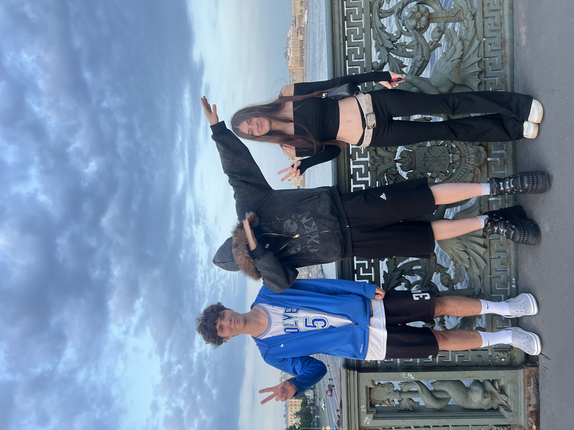 Three young people stand on a bridge. The person on the left is a male wearing a blue zip-up jacket over a white jersey with the number 5, black shorts, white socks, and grey Crocs. He is making a peace sign with his left hand. In the center, a person in a dark hooded jacket with fur trim, black shorts, and platform shoes is obscuring their face with their arm, performing a dab pose. To the right, a female is wearing a black off-the-shoulder crop top, black pants, a light-colored belt, and white sneakers. She is also making a peace sign with her left hand. The background shows a body of water, a cityscape with buildings, and a cloudy sky. The bridge has ornate metal railings with decorative elements.