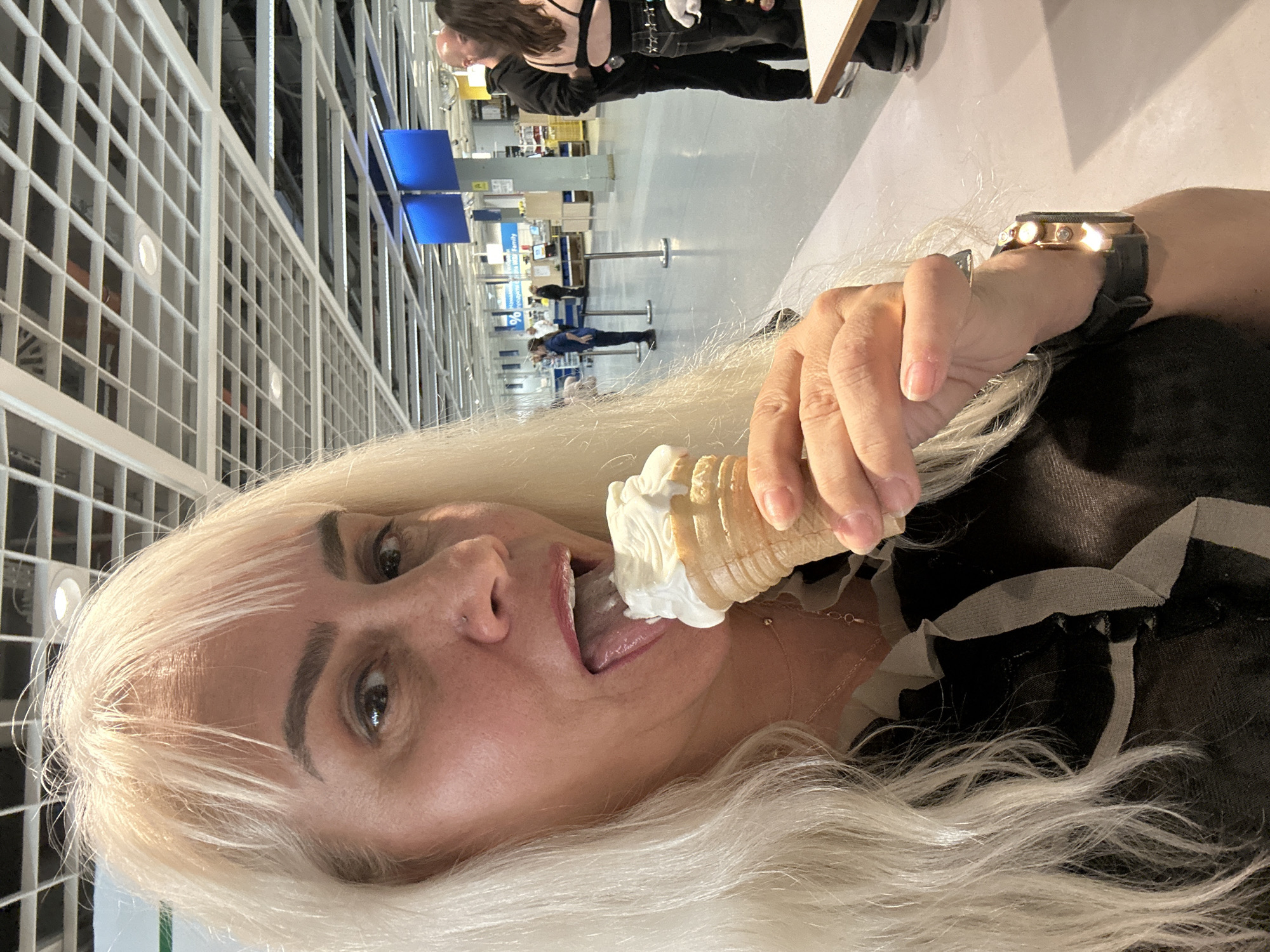 A woman with blonde hair is eating an ice cream cone in what appears to be a large, open commercial space, possibly a store. In the background, a grid-like structure spans the ceiling, suggesting a building interior. People are walking in the distance, and there are signs visible, though unreadable due to the distance. The person taking the picture is at Ikea, Sofia, Bulgaria. The focus is on the woman in the foreground, emphasizing her action of eating the ice cream. The person taking the image has a watch on.