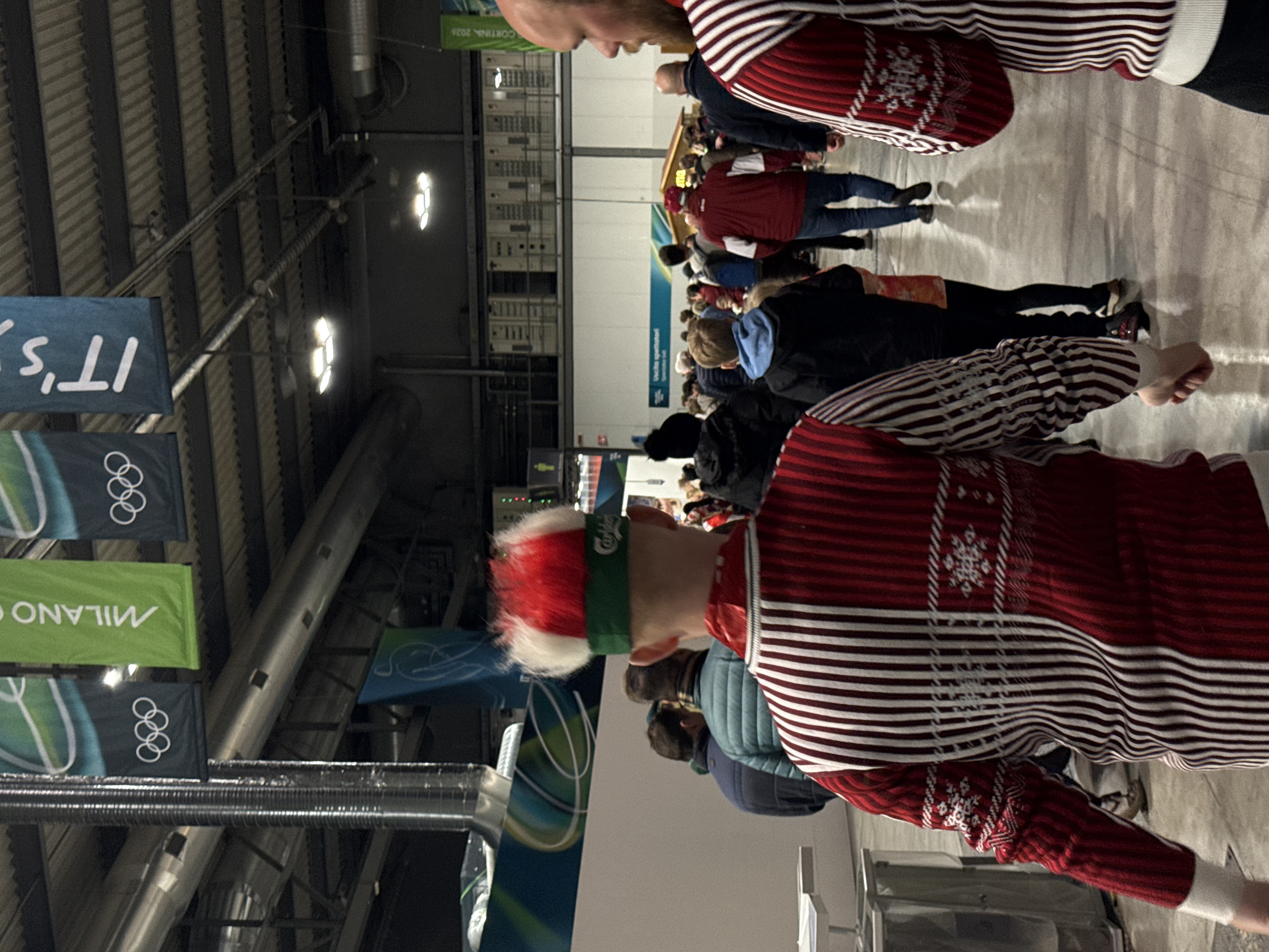 A crowd of people stands indoors, many wearing winter attire. Dominating the foreground are two individuals in red and white striped sweaters. One wears a red, white, and green hat. Olympic banners with "Milano Cortina 2026" hang from the ceiling, alongside banners that read "It's." The setting appears to be an indoor event or gathering.