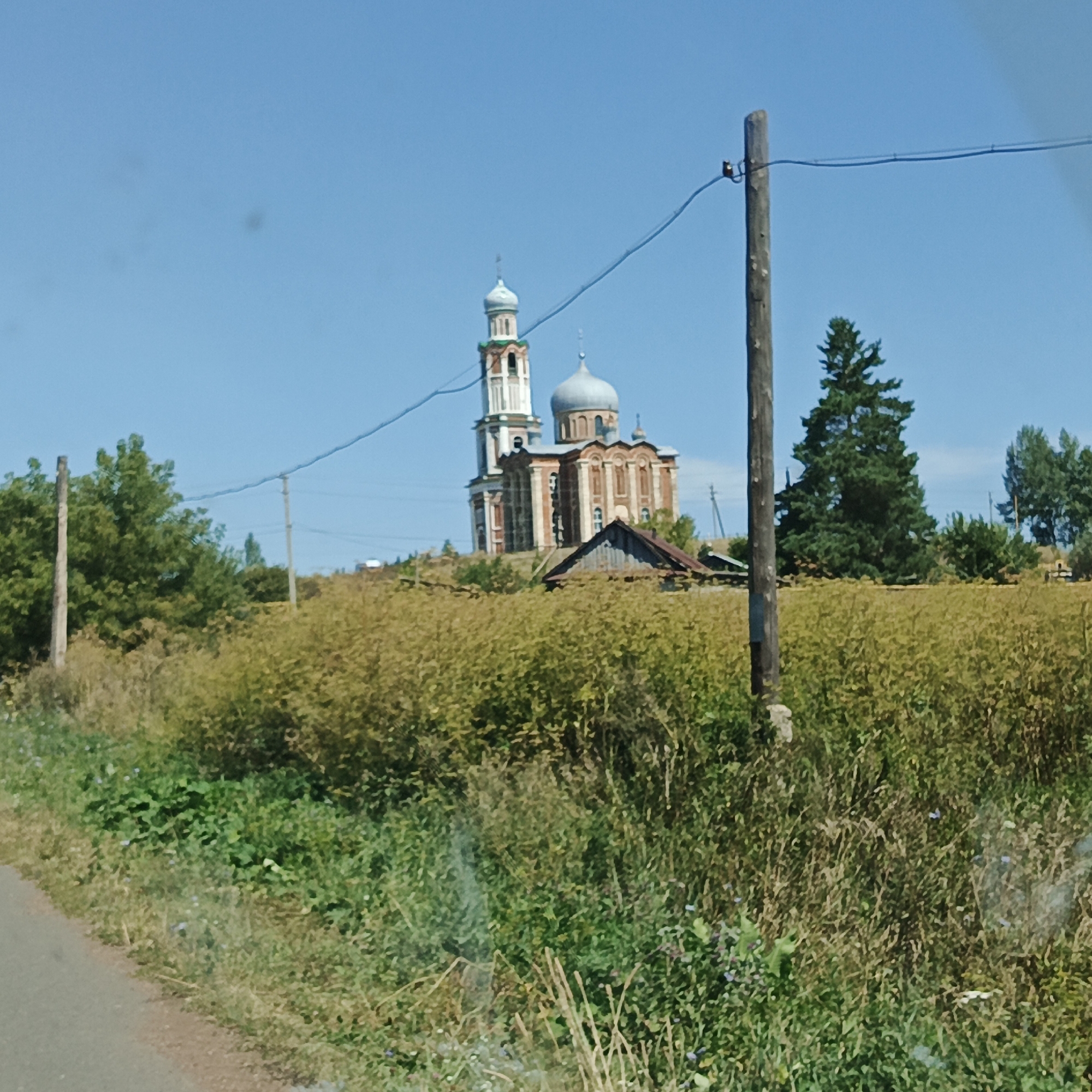The image captures a rural landscape under a clear blue sky, possibly in the village of Tugustemir, Russia, given the context. In the background, a prominent church with multiple domes and a bell tower stands on a slight elevation. The church's architecture appears to be traditional Russian Orthodox, with red brick and white decorative elements. In front of the church, a small, rustic wooden structure with a dark roof is visible.
The foreground is dominated by a field of dry, yellowish-brown grass and some green vegetation. A dirt or asphalt road runs along the left edge of the frame. A wooden utility pole with wires stretches diagonally across the upper portion of the image. Several trees, including evergreens and deciduous trees, are scattered throughout the scene, with a dense cluster of green trees to the left and a solitary tall evergreen to the right of the church. The overall impression is one of a serene, sunlit, rural setting. There are no people or animals visible in the image.