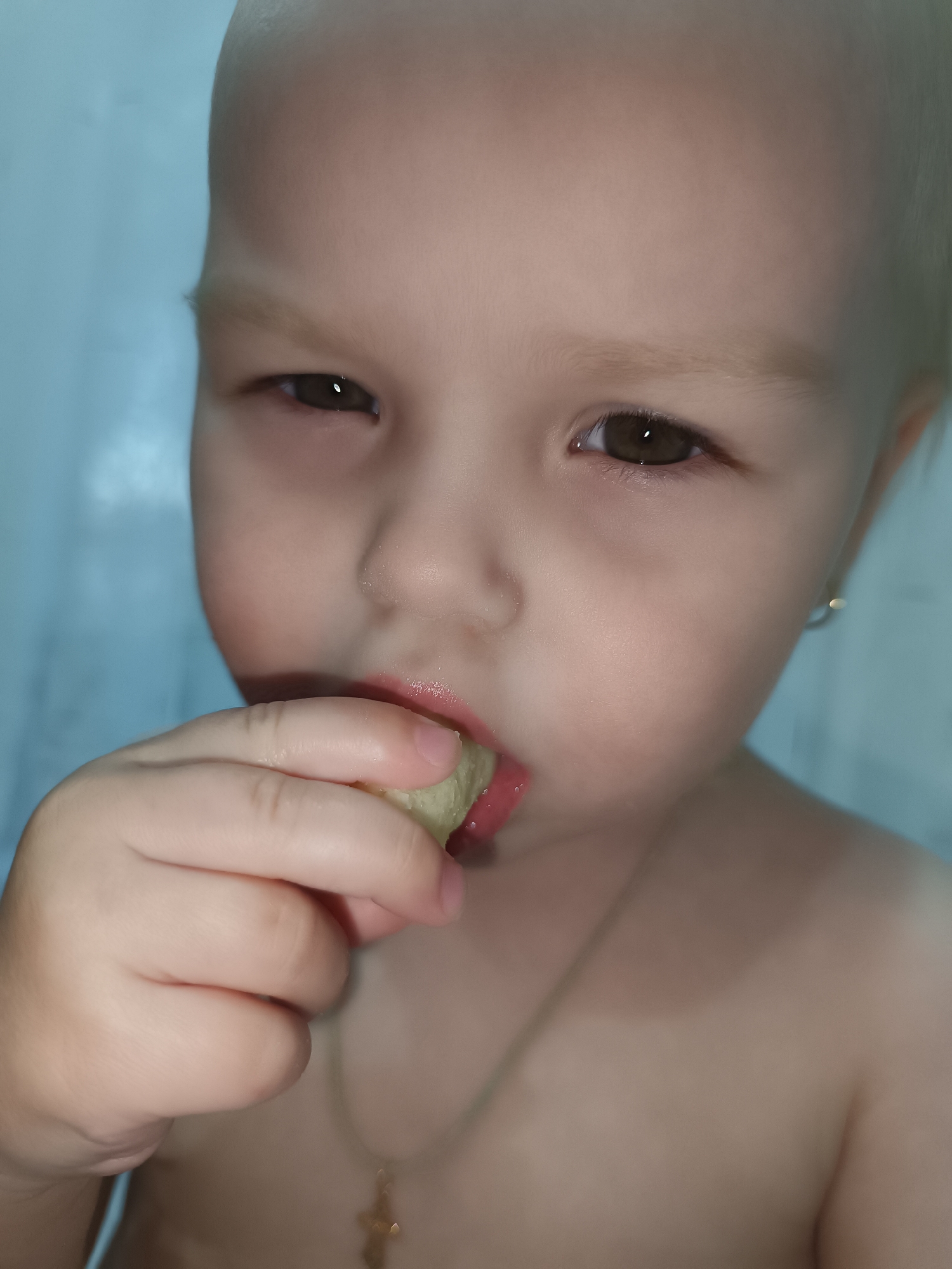 A close-up shot shows a young child eating something held in their left hand. The child has light skin, light hair, and brown eyes. A small gold earring is visible in their left ear. They are wearing a gold necklace with a small cross pendant. The background appears to be a light blue wall. The location is Chaikovsky, Russia.