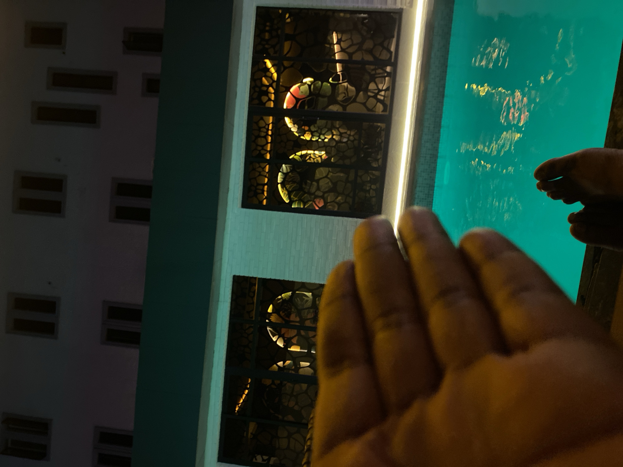 A nighttime photograph shows a person's hand in the foreground, obscuring the view. Beyond the hand is a building with distinctive, decorative windows lit from within. The building appears to be adjacent to a turquoise-colored swimming pool. A person's toes can be seen near the edge of the pool. The location is Kubwa, Nigeria.