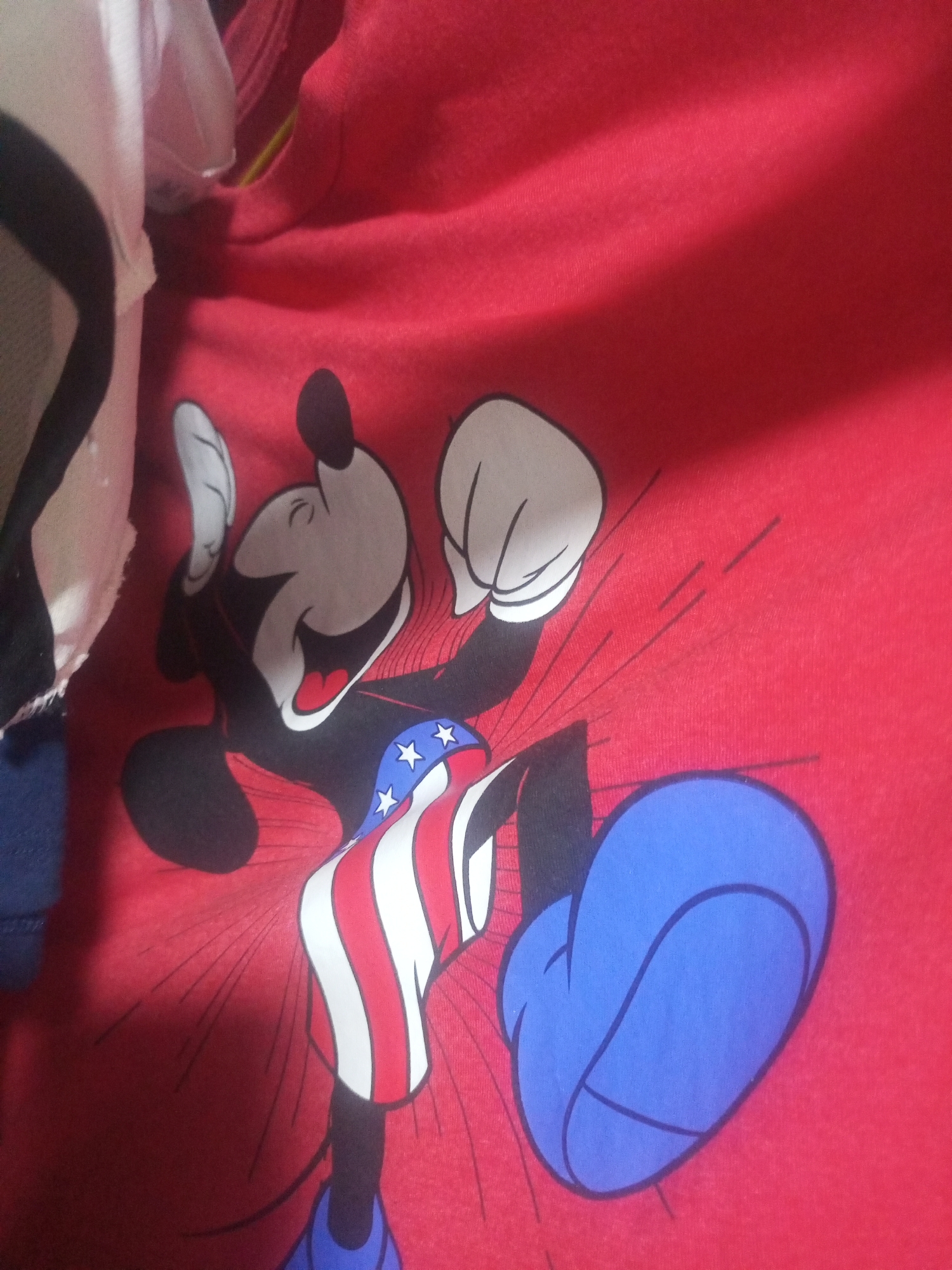The image shows a close-up view of a red fabric with a graphic of Mickey Mouse. Mickey Mouse is depicted with a wide, open mouth and its arms are raised. It is wearing shorts with red and white stripes and a pattern of blue stars on a white band. The character's feet are depicted as large blue shoes. The fabric is wrinkled, suggesting it is a piece of clothing. There are some indistinct white and dark objects visible in the upper left portion of the image, possibly part of other clothing items. The setting is not specified as being in La Tagua, Venezuela; no urban or geographical context is provided.