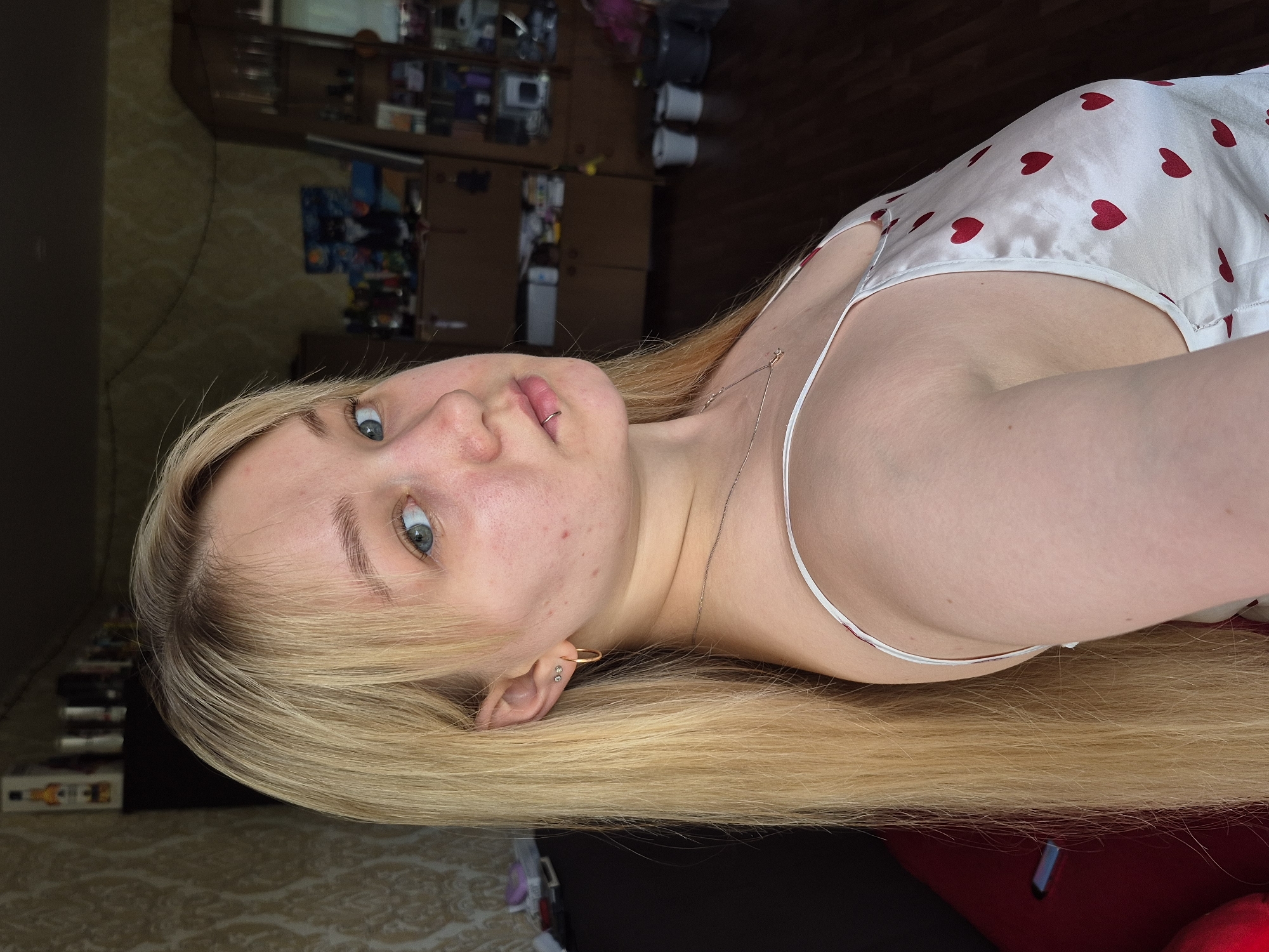 A young Caucasian woman with long blonde hair and blue eyes is depicted indoors. She is wearing a white top with red heart patterns. She has fair skin with some blemishes visible on her face and a lip piercing. She also has gold hoop earrings. A thin necklace is visible around her neck. The background appears to be a cluttered domestic setting with furniture and various objects. The location may be Chaikovsky, Russia, but this cannot be confirmed from the image alone.